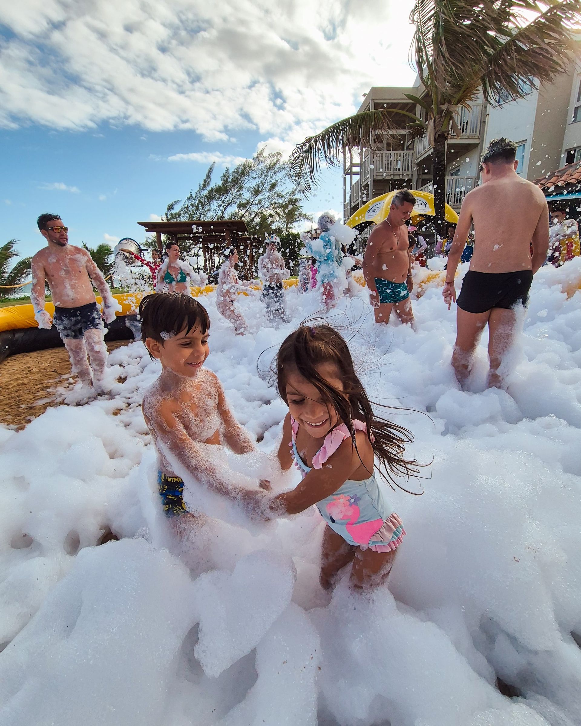 Crianças brincando em uma piscina externa cheia de espuma perto de um resort, com adultos ao fundo.