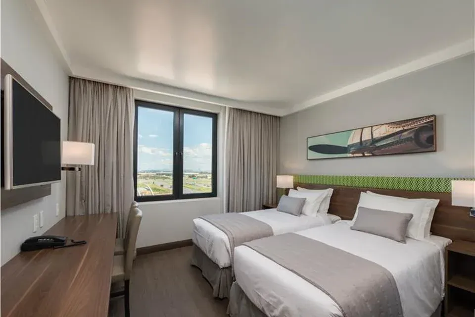 Quarto de hotel moderno com duas camas de solteiro, vista para a janela, escrivaninha e TV de parede.