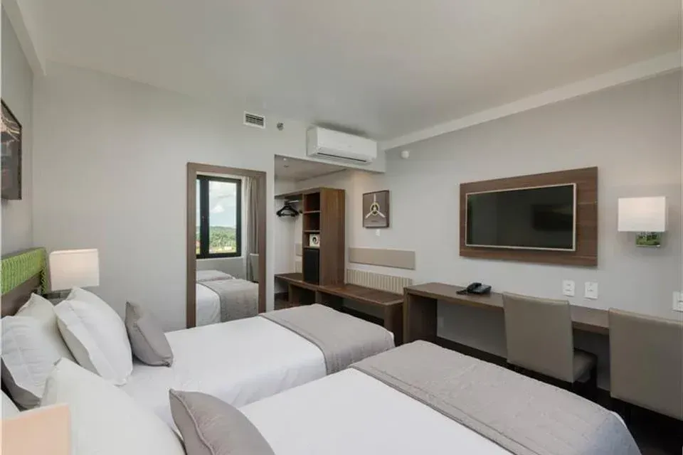 Quarto de hotel moderno com duas camas de solteiro, TV de parede, escrivaninha, espelho e ar-condicionado.