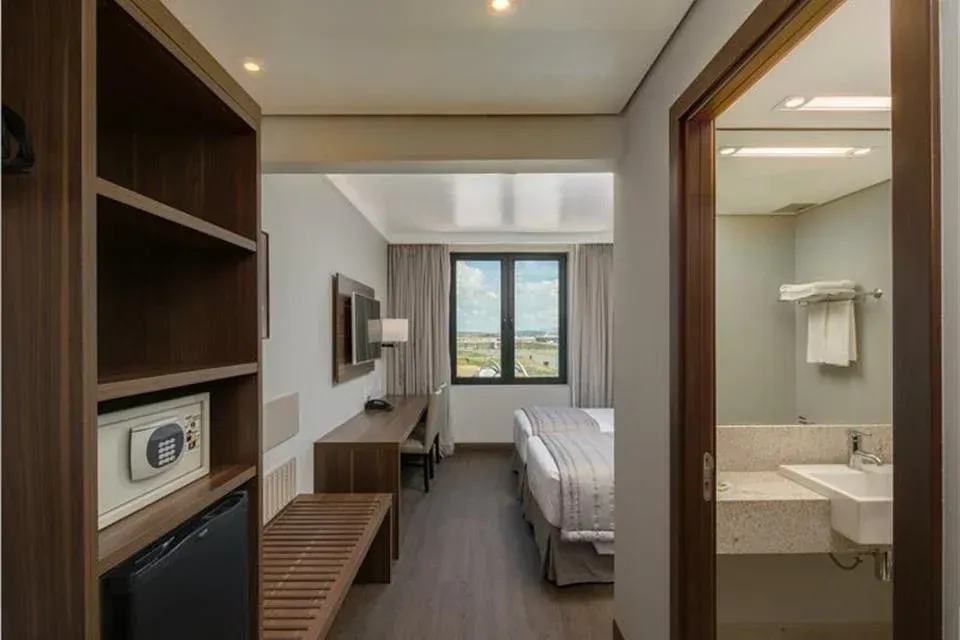 Quarto de hotel compacto com duas camas de solteiro, escrivaninha, janela e banheiro privativo visível pela porta.