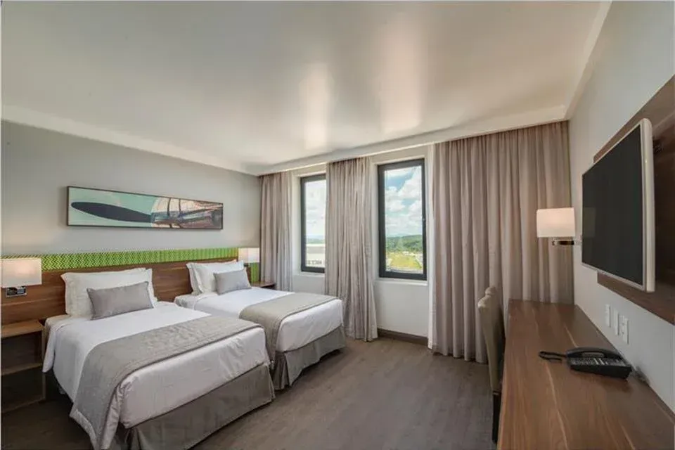 Quarto de hotel moderno com duas camas, janela grande, TV de parede e cortinas bege.