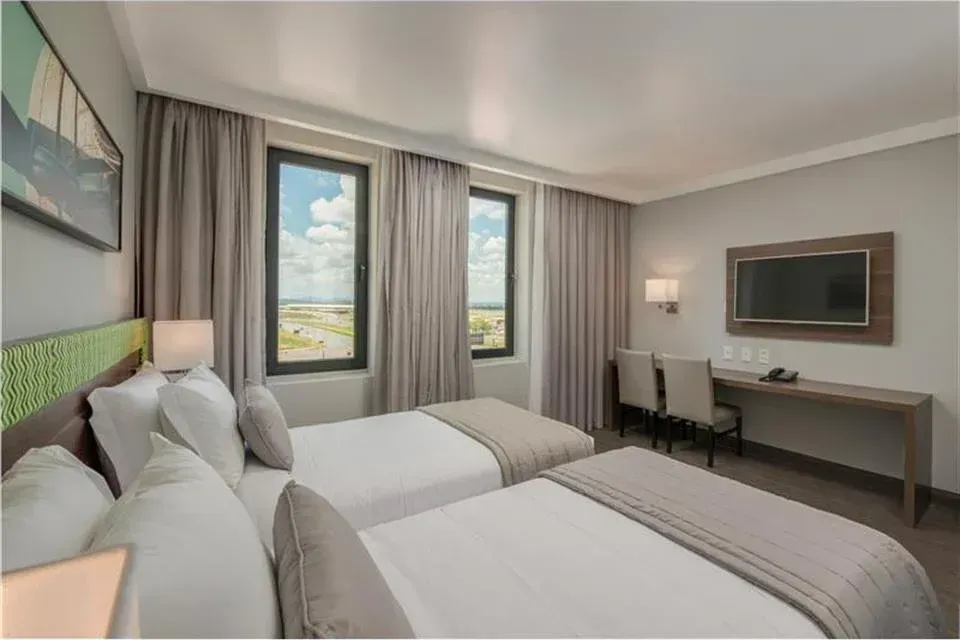 Quarto de hotel moderno com duas camas de solteiro, escrivaninha, TV de parede e janelas com vista para a cidade.