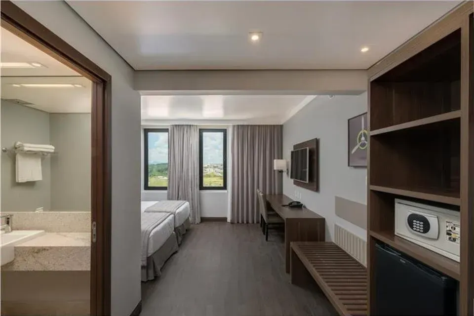 Quarto de hotel moderno com duas camas, escrivaninha, TV de parede e banheiro visível através de uma porta aberta.