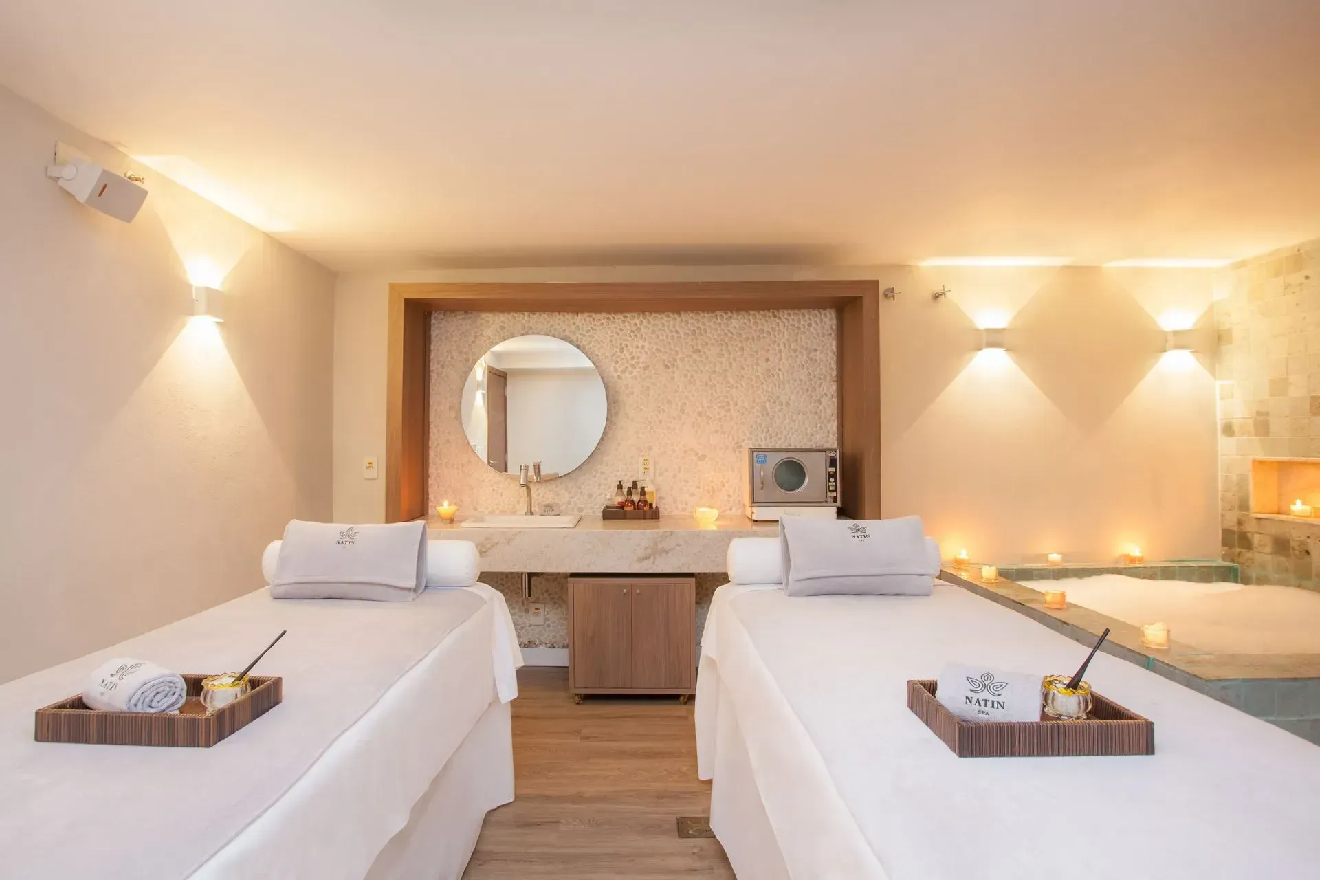 Sala de tratamento de spa com duas camas de massagem, iluminação suave e espelho de maquilhagem.