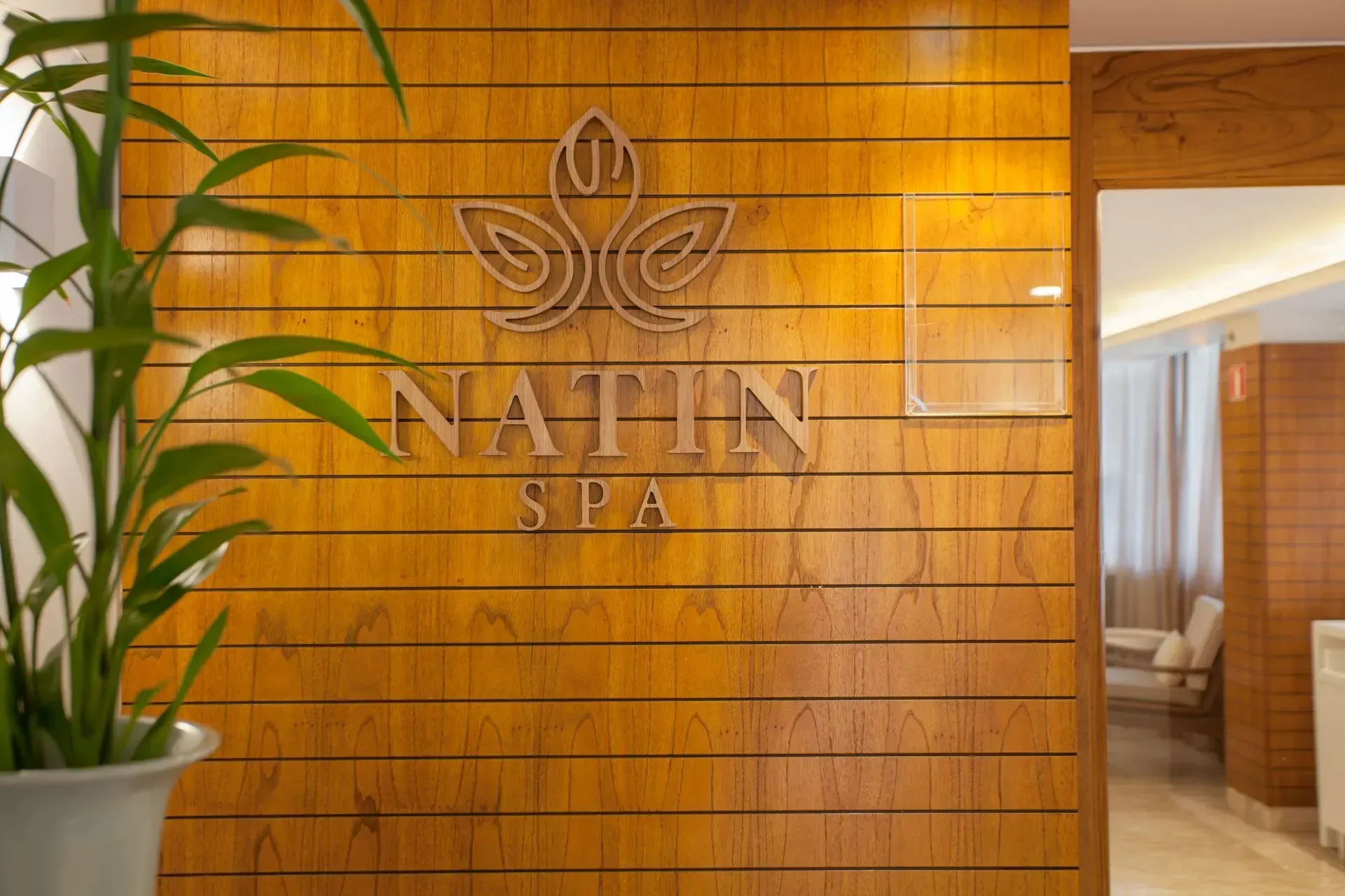 Logotipo do Natin Spa em uma parede de madeira aconchegante, com uma planta e um corredor de entrada com iluminação suave nas proximidades.