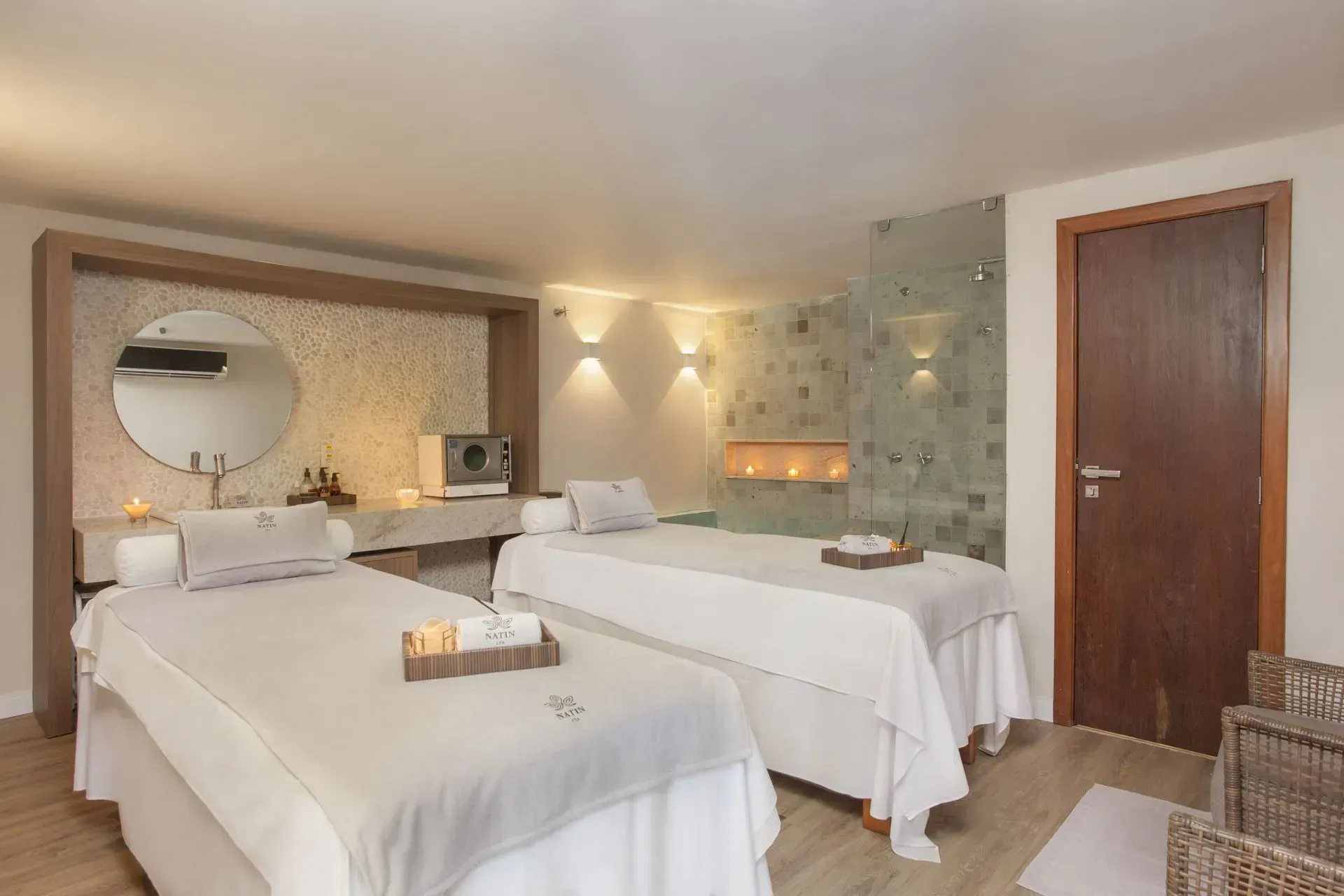 Quarto de hotel luminoso com duas camas de solteiro, espelho redondo, portas de madeira e iluminação suave nas paredes.