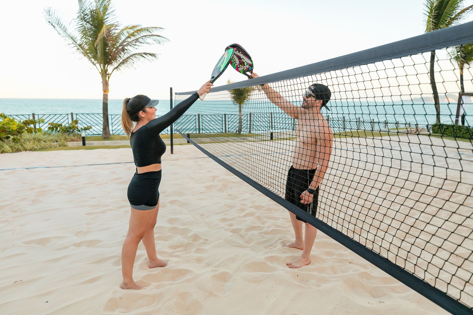 Duas pessoas ajustando uma rede de vôlei em uma praia de areia perto de palmeiras e do oceano.