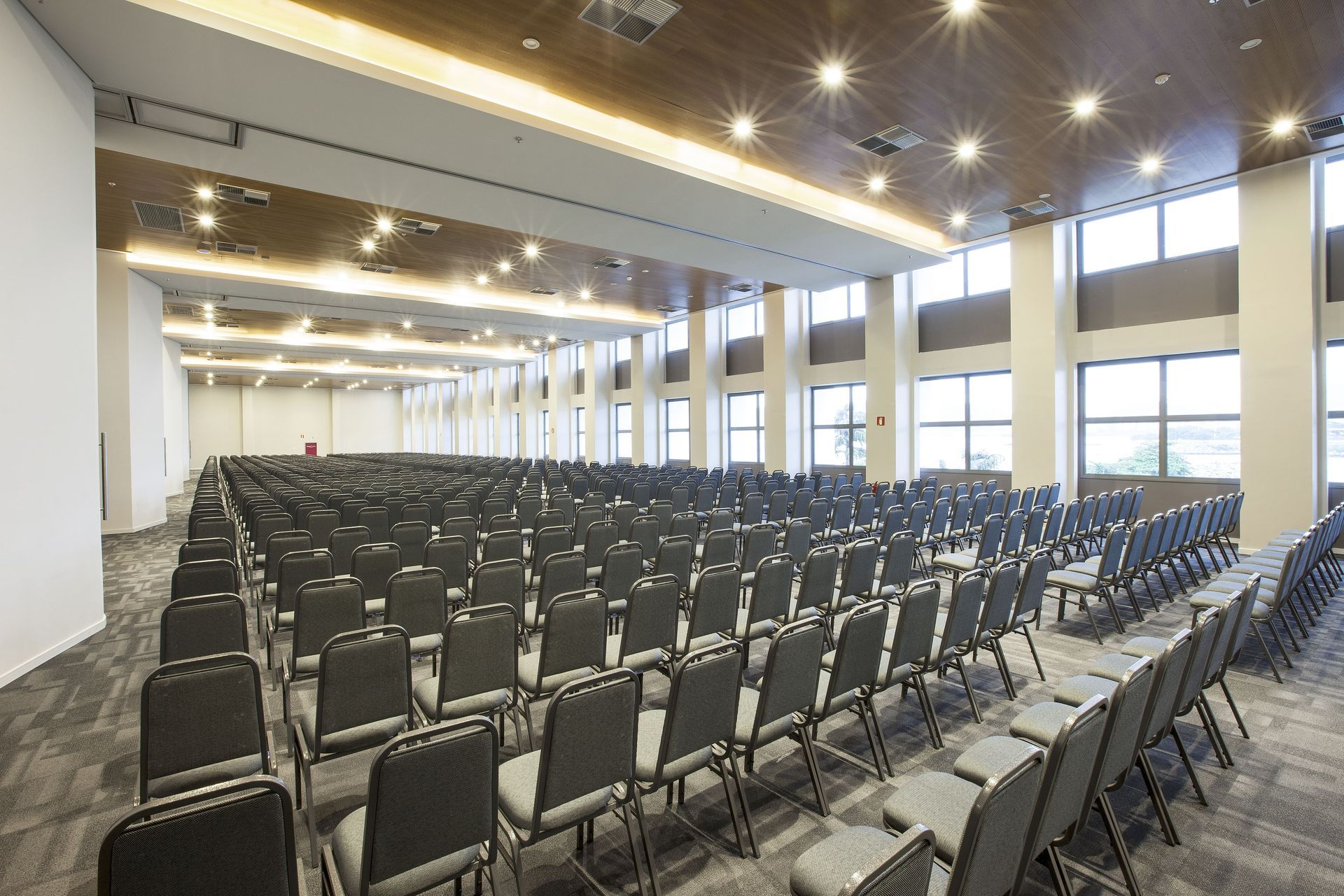 Sala de conferências vazia com fileiras de cadeiras pretas, grandes janelas e luzes no teto.