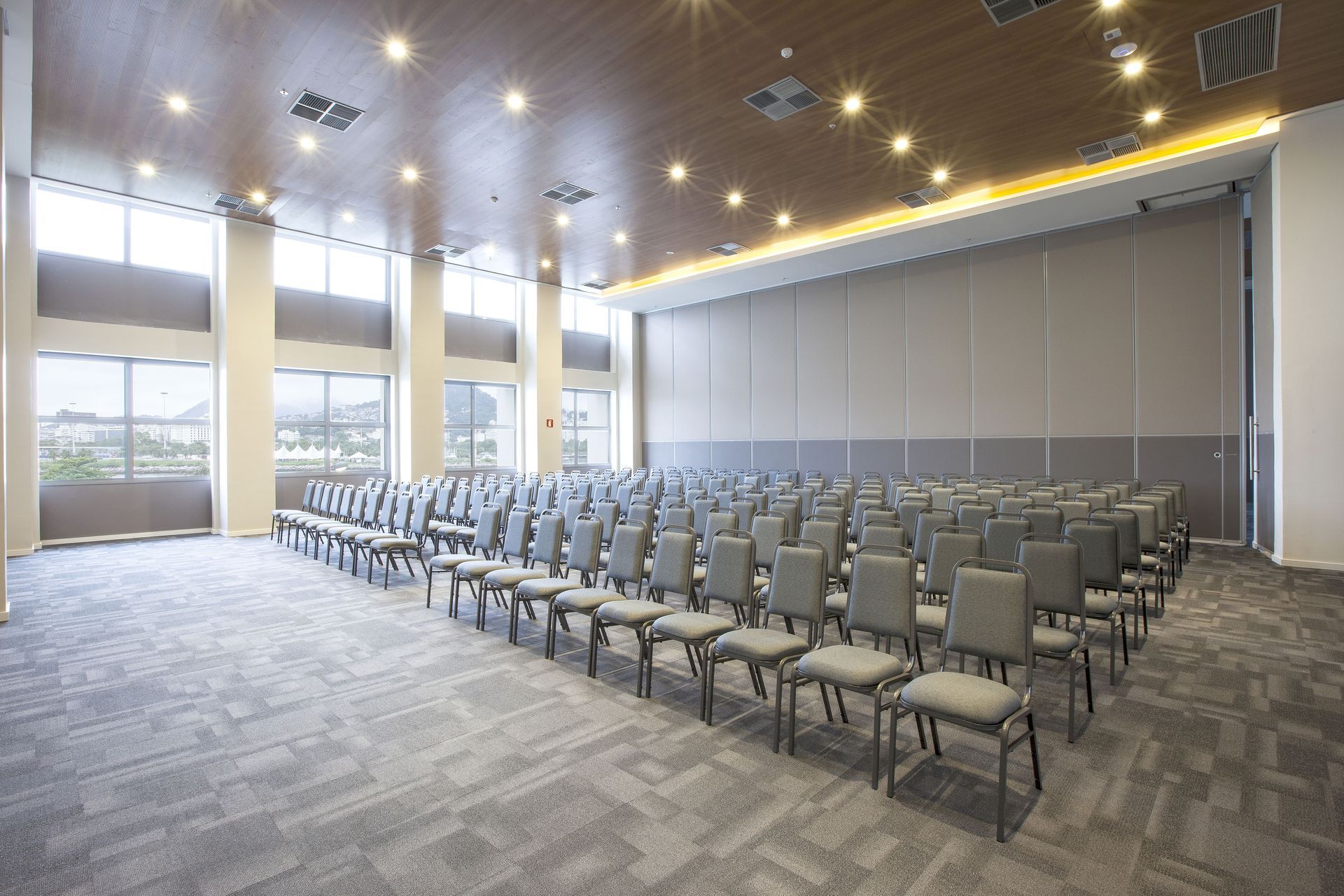 Sala de conferências vazia com fileiras de cadeiras cinzentas e grandes janelas ao longo de uma das paredes.
