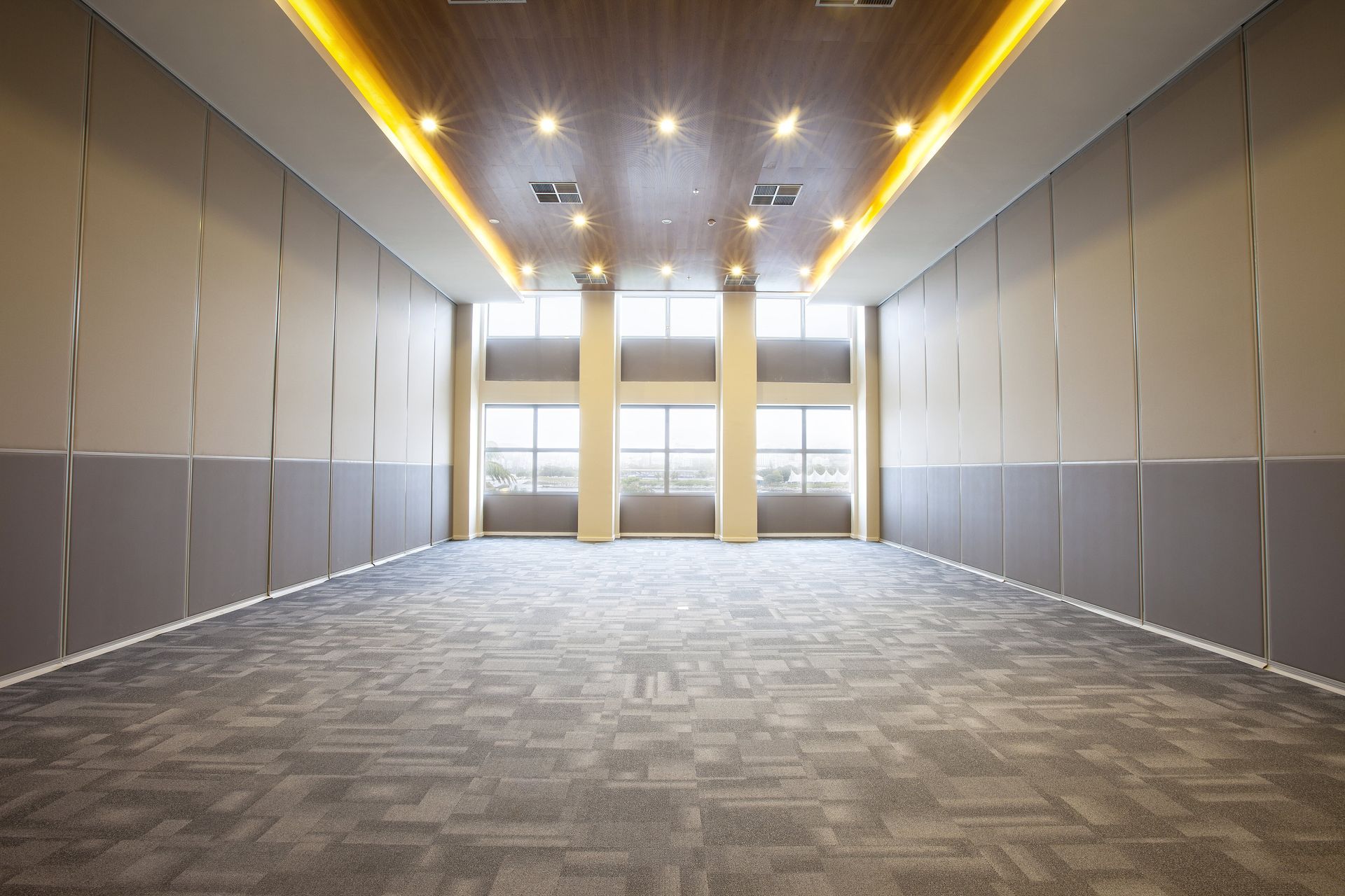 Sala de conferências moderna e vazia, com piso acarpetado, iluminação embutida no teto e grandes janelas na extremidade oposta.