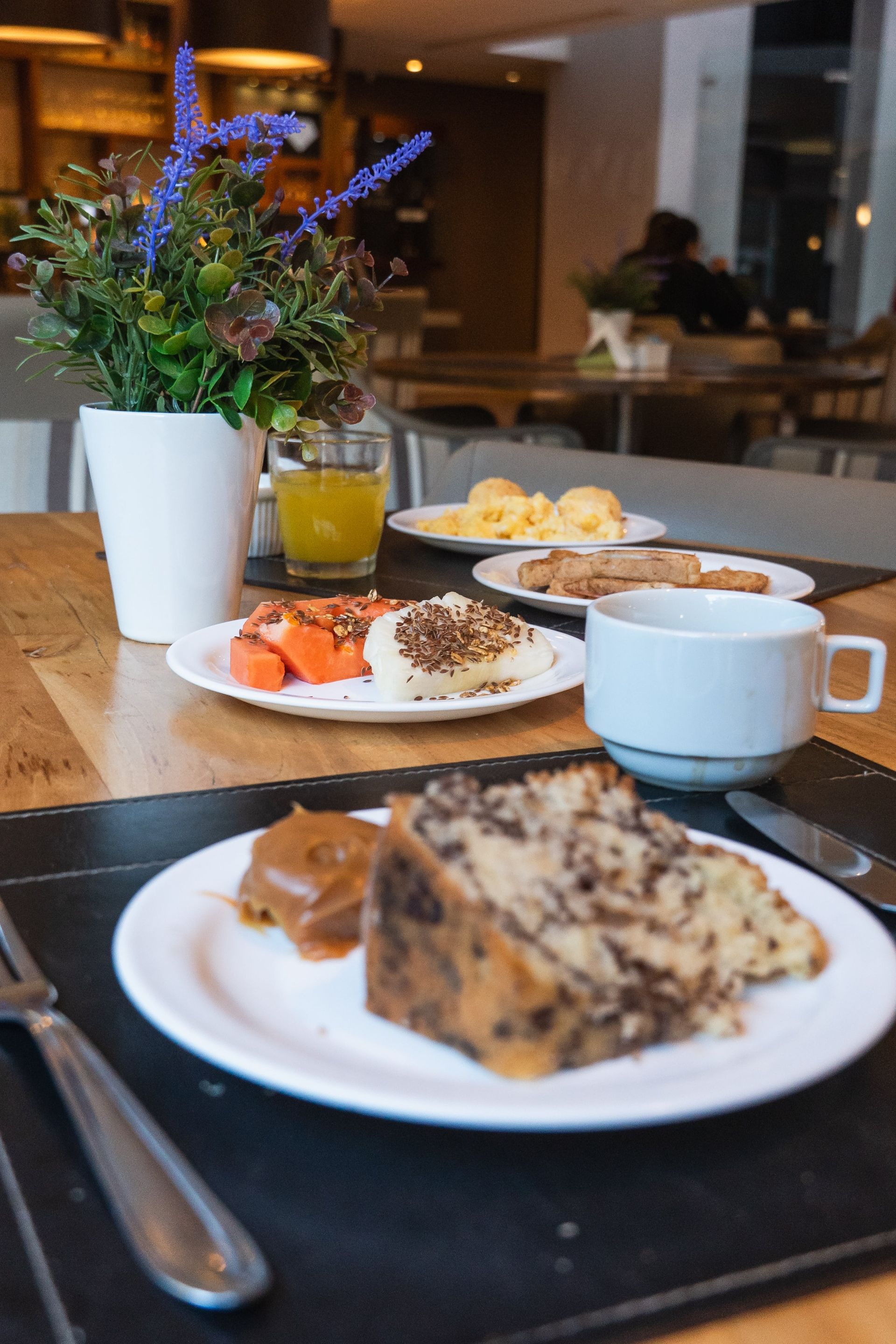 Mesa de café da manhã com doces, frutas, café e um vaso de flores roxas em um café.