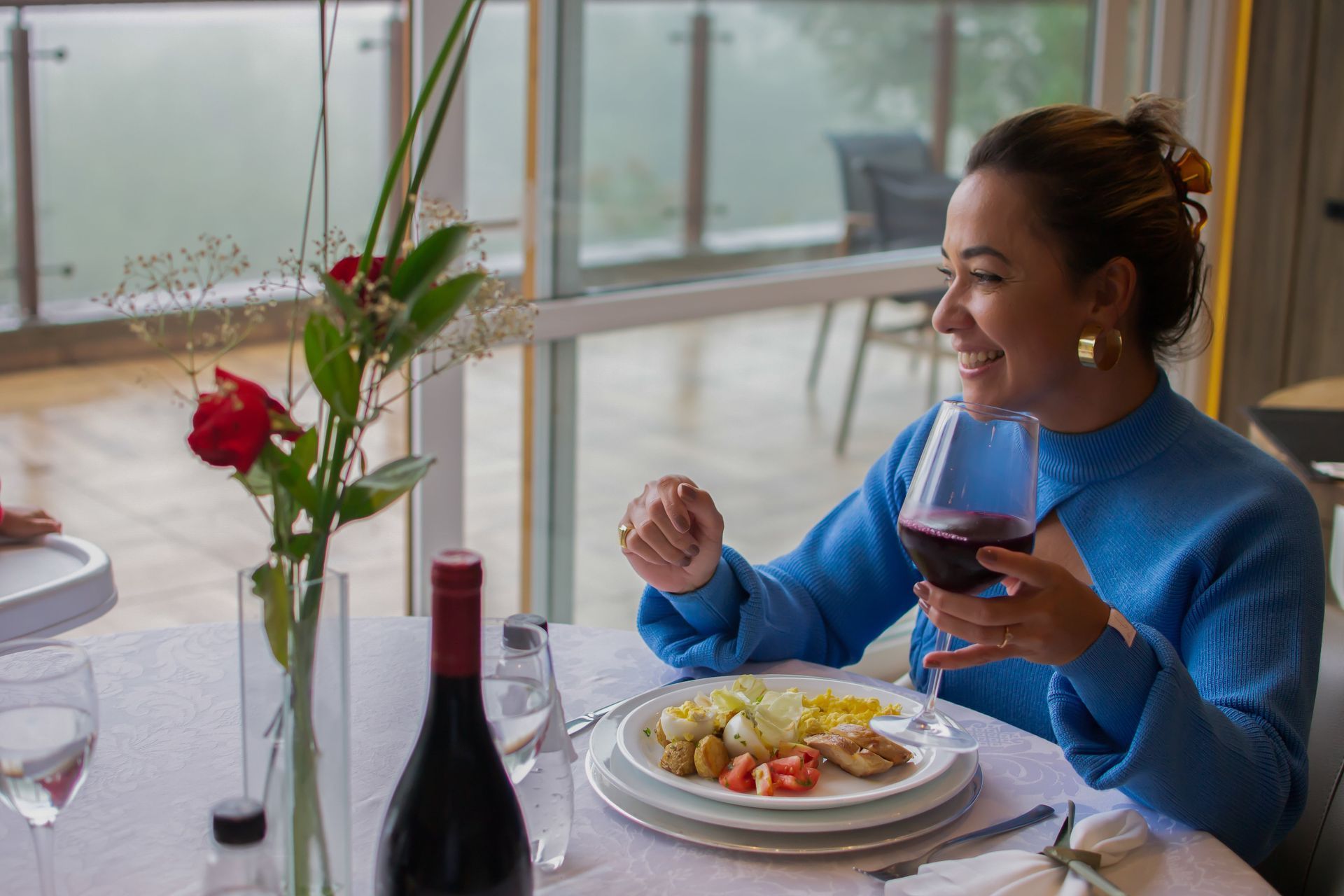 Pessoa jantando em uma mesa perto da janela, segurando uma taça de vinho e sorrindo, com um prato de comida e flores por perto.