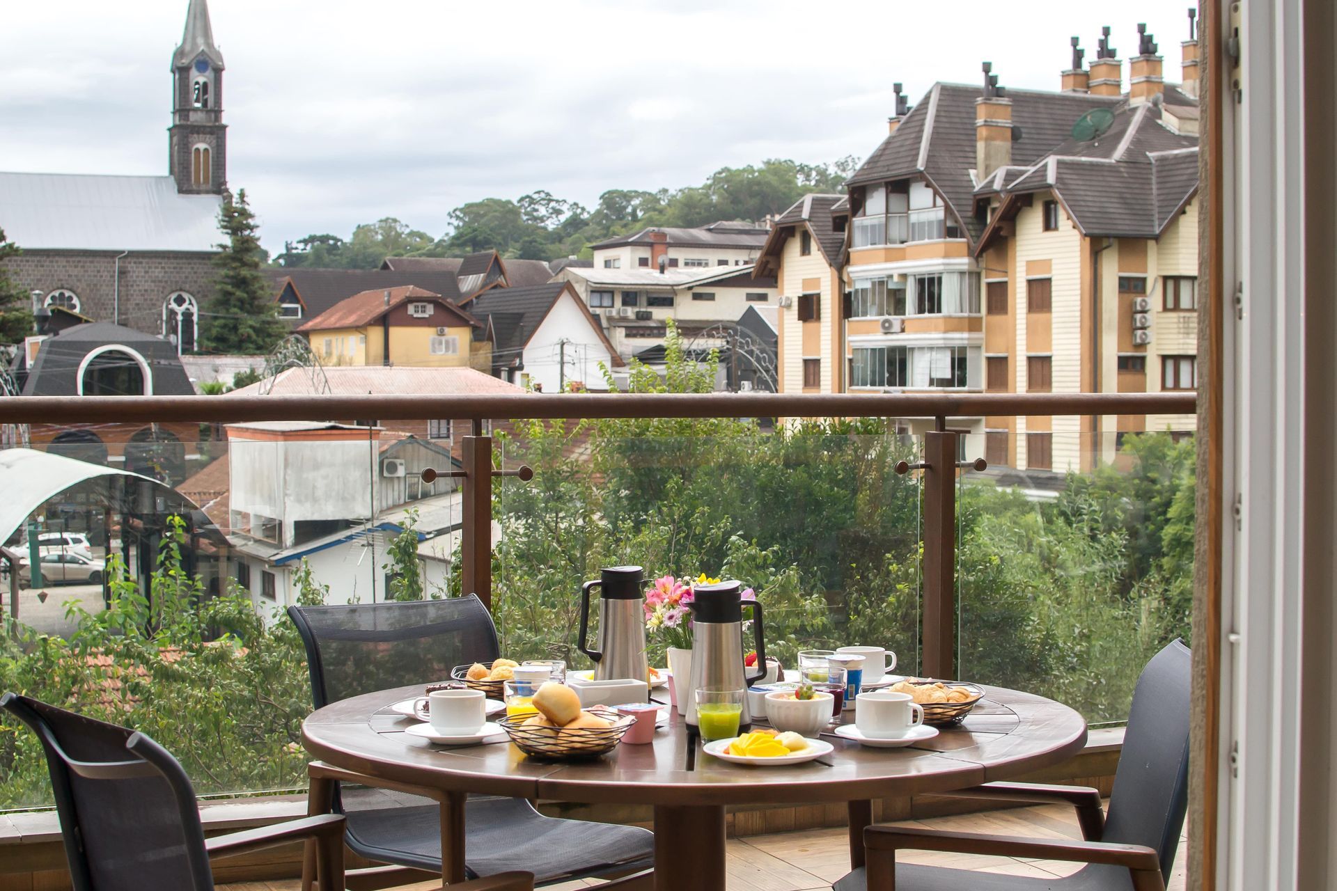 Mesa de café da manhã na varanda com chá, frutas e doces, com vista para uma vila de casas e a torre de uma igreja.