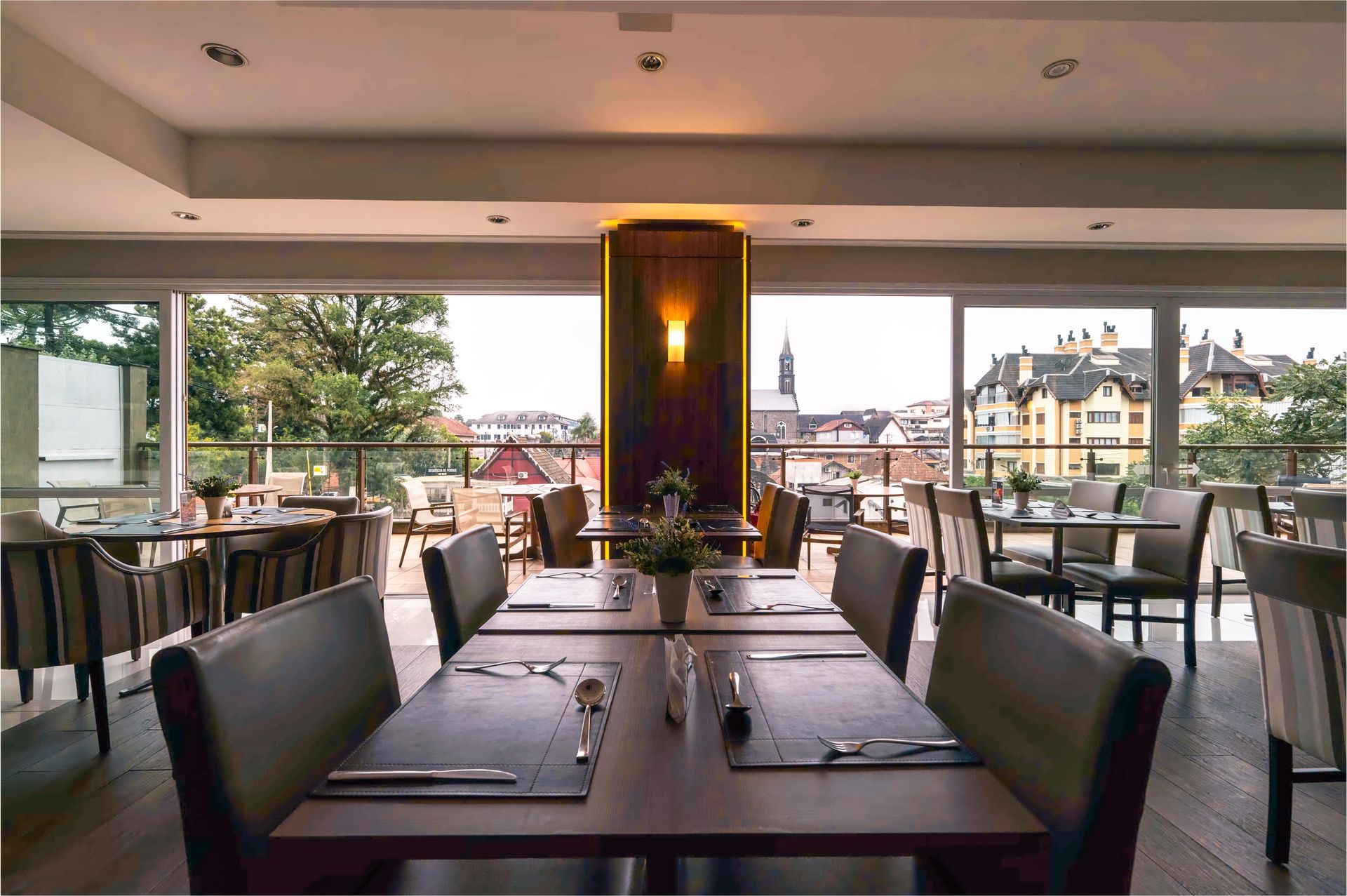 Sala de jantar moderna do restaurante com mesas postas, cadeiras pretas e vista para a cidade através de grandes janelas.