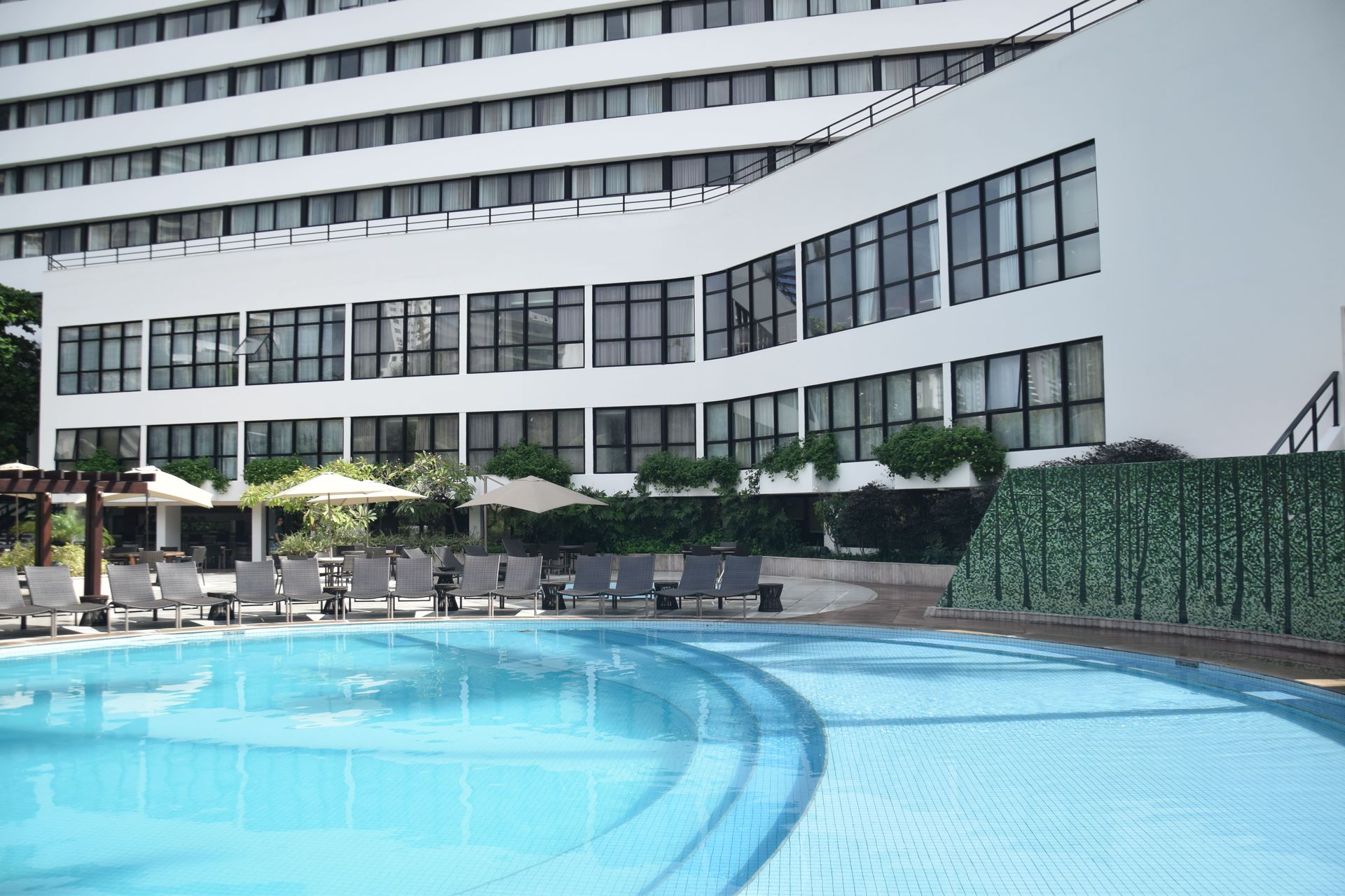 Piscina externa do hotel ao lado de um prédio branco moderno, com espreguiçadeiras e guarda-sóis.