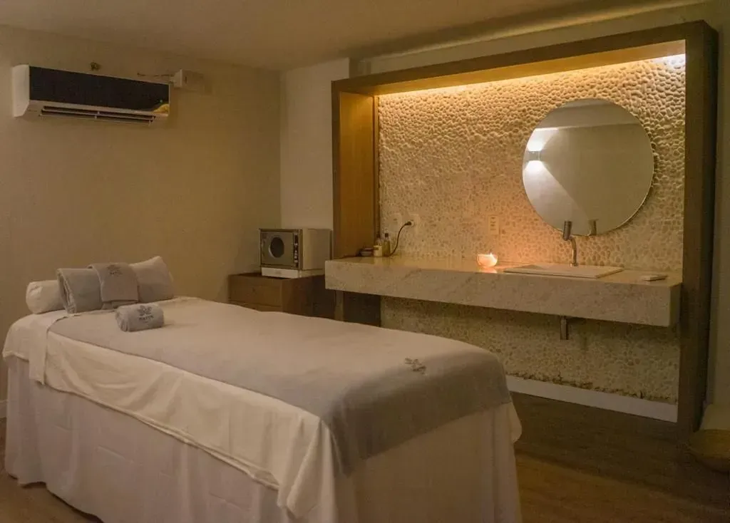 Sala de tratamento de spa com maca de massagem, espelho redondo e parede com textura aconchegante.