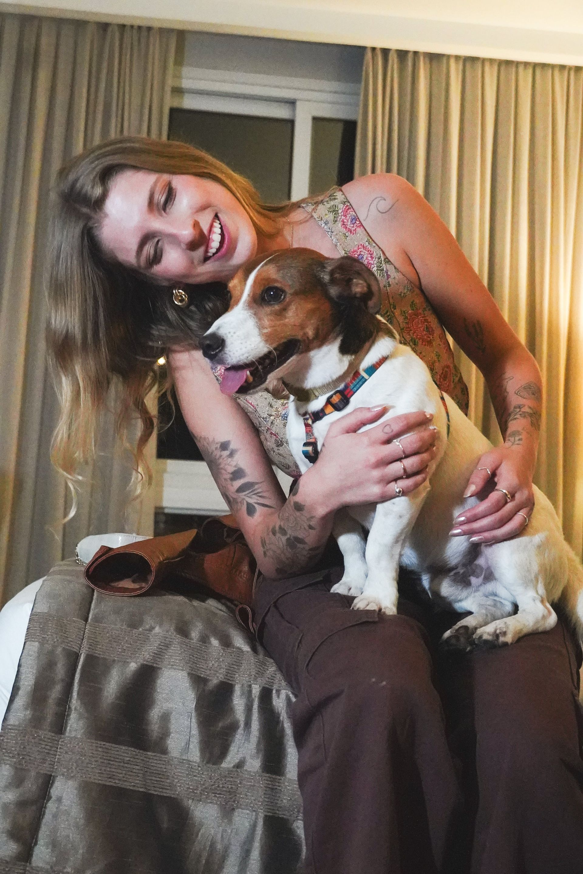 Mulher sentada dentro de casa, sorrindo e segurando um pequeno cachorro marrom e branco no colo, perto de uma janela.