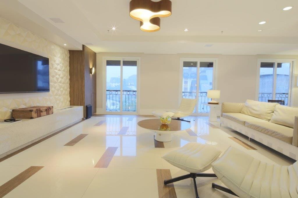 Sala de estar moderna e iluminada com sofás brancos, piso brilhante, TV de parede e grandes janelas com vista para a cidade.
