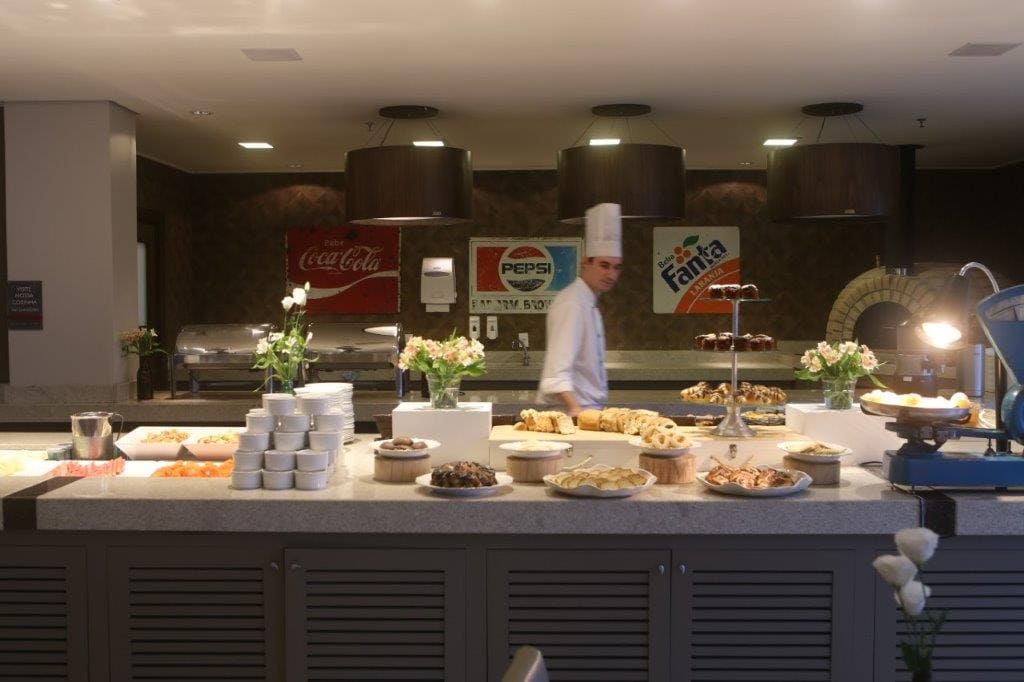 Balcão de buffet com bandejas de comida na área de refeições de um hotel, com um chef em pé atrás dele.