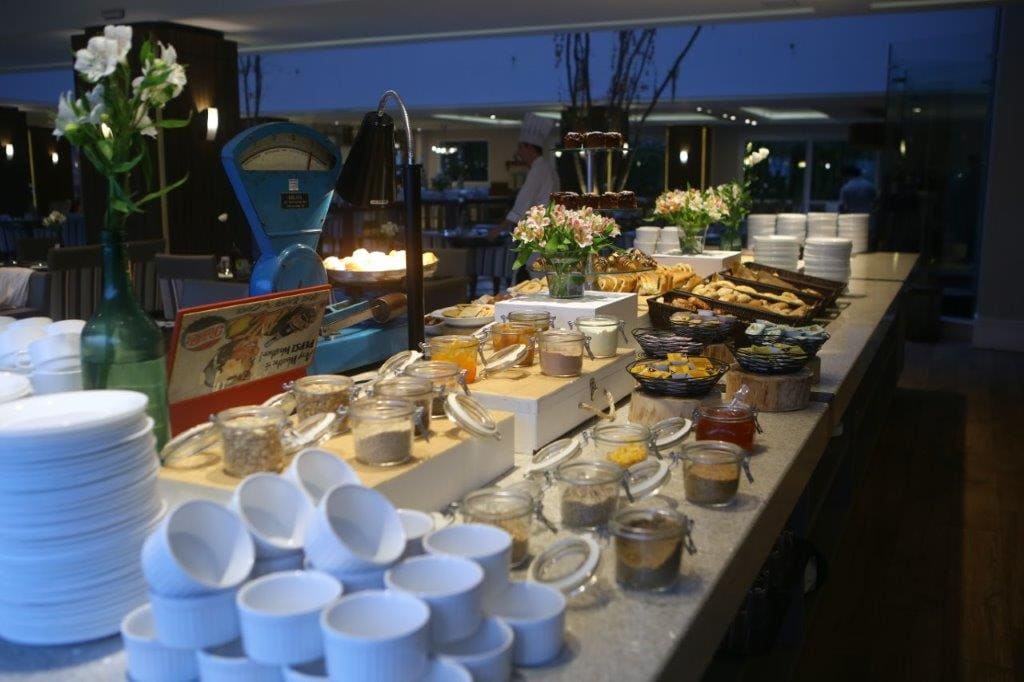 Mesa de buffet com xícaras, pratos e bandejas de comida variadas empilhadas em um ambiente interno com iluminação suave.