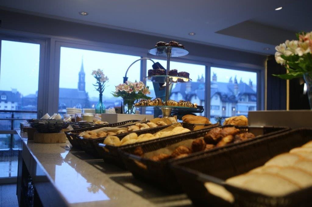 Mesa de buffet com doces e bandejas de sobremesas junto a grandes janelas com vista para a paisagem urbana.