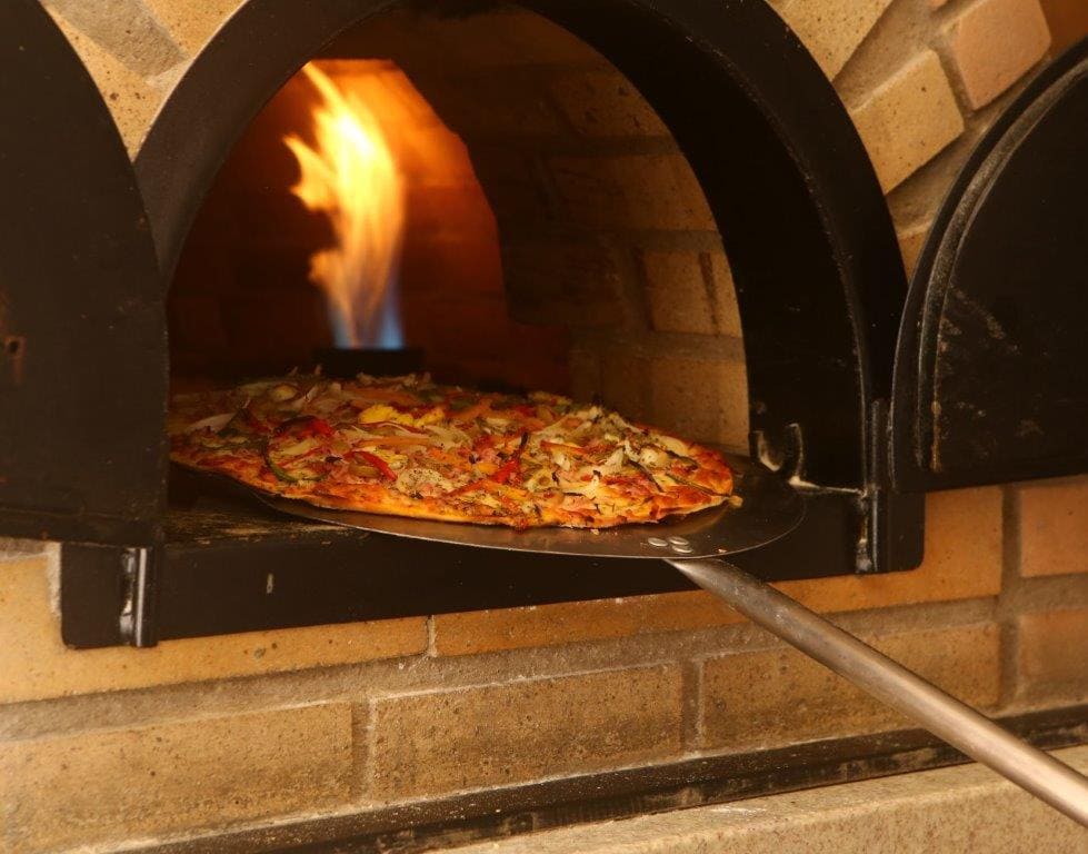 Pizza assando em forno de tijolos a lenha, com chamas atrás e uma pá de metal como apoio.