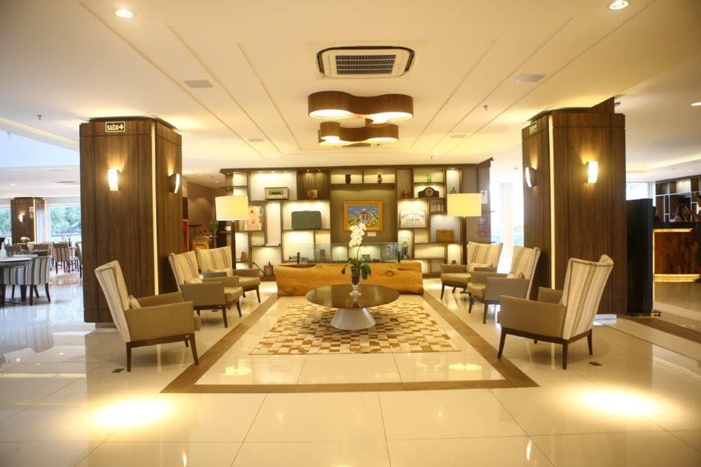 Lobby de hotel moderno com assentos bege, piso brilhante e iluminação aconchegante ao redor de uma mesa de exposição central.