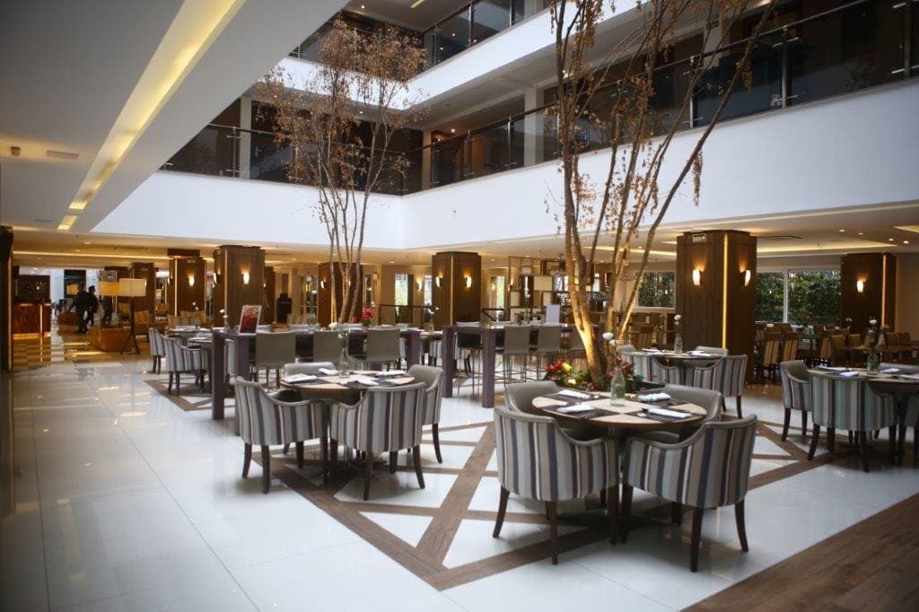 Lobby de hotel moderno com mesas de jantar redondas, cadeiras e altos galhos decorativos sob iluminação aconchegante.