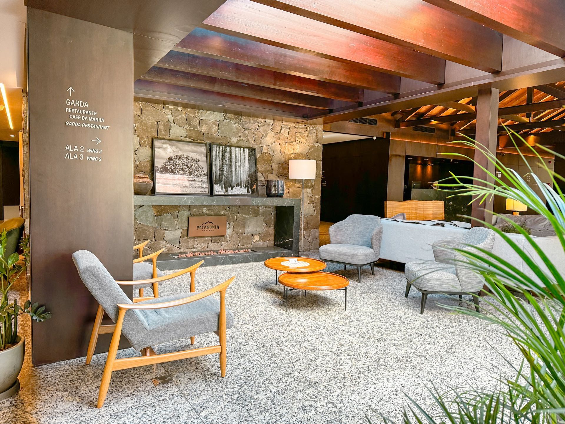 Sala de estar rústica e aconchegante com cadeiras cinza, mesa laranja, vigas de madeira e lareira de pedra no interior.