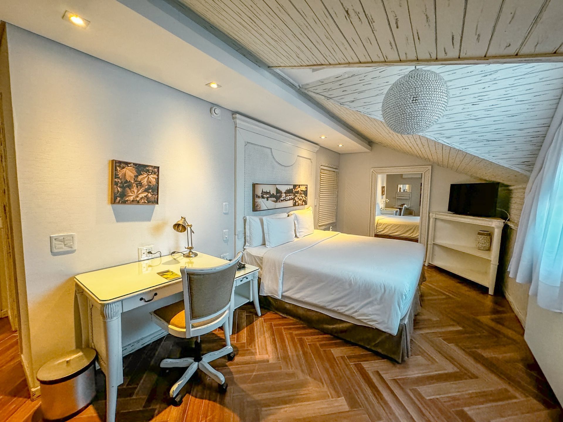 Quarto luminoso no sótão com cama, escrivaninha, cadeira e piso de madeira com padrões, sob um teto inclinado com claraboias.