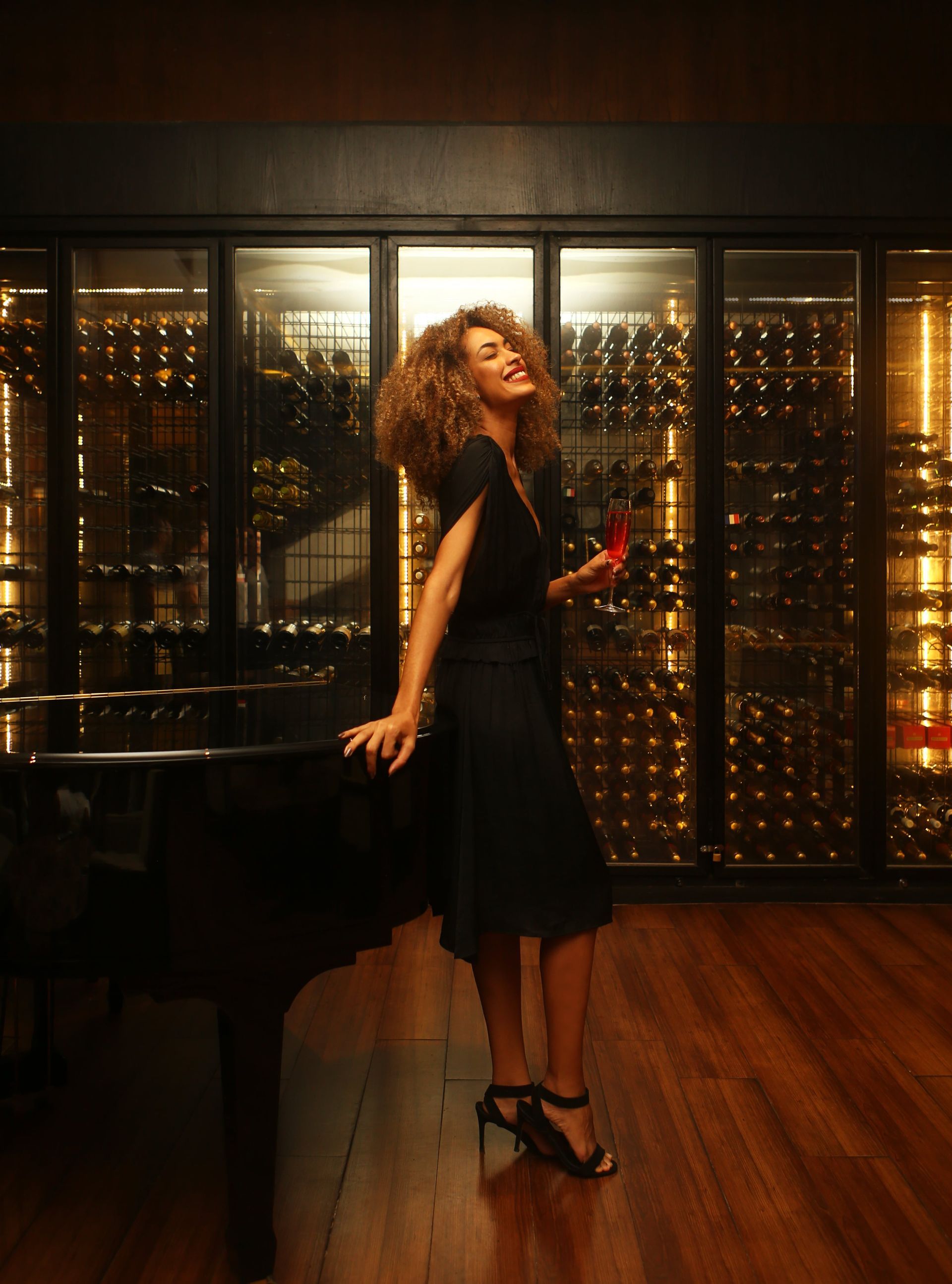 Mulher de vestido preto posando em uma adega pouco iluminada com luz ambiente quente.