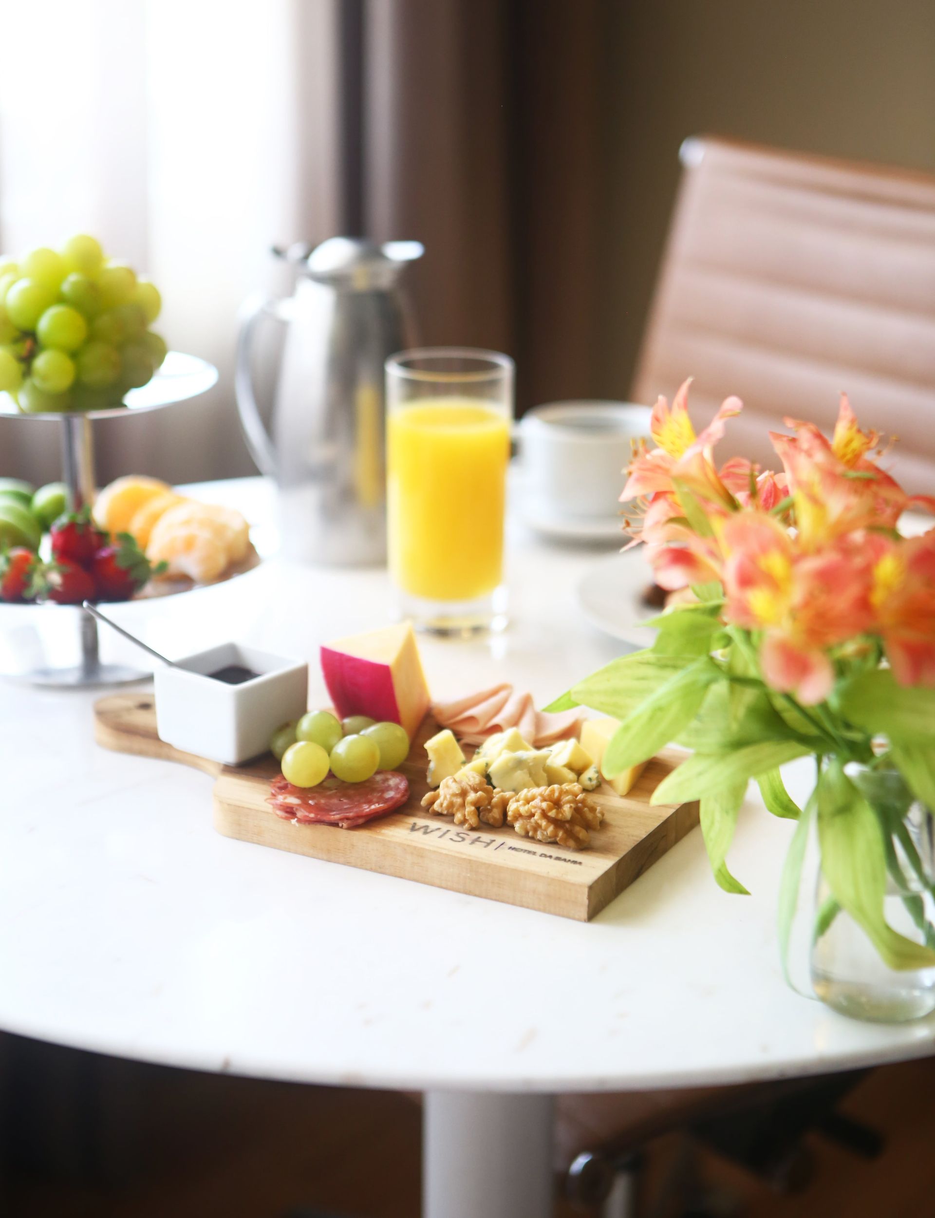 Buffet de café da manhã com frutas, sucos, doces e flores sobre uma mesa branca.