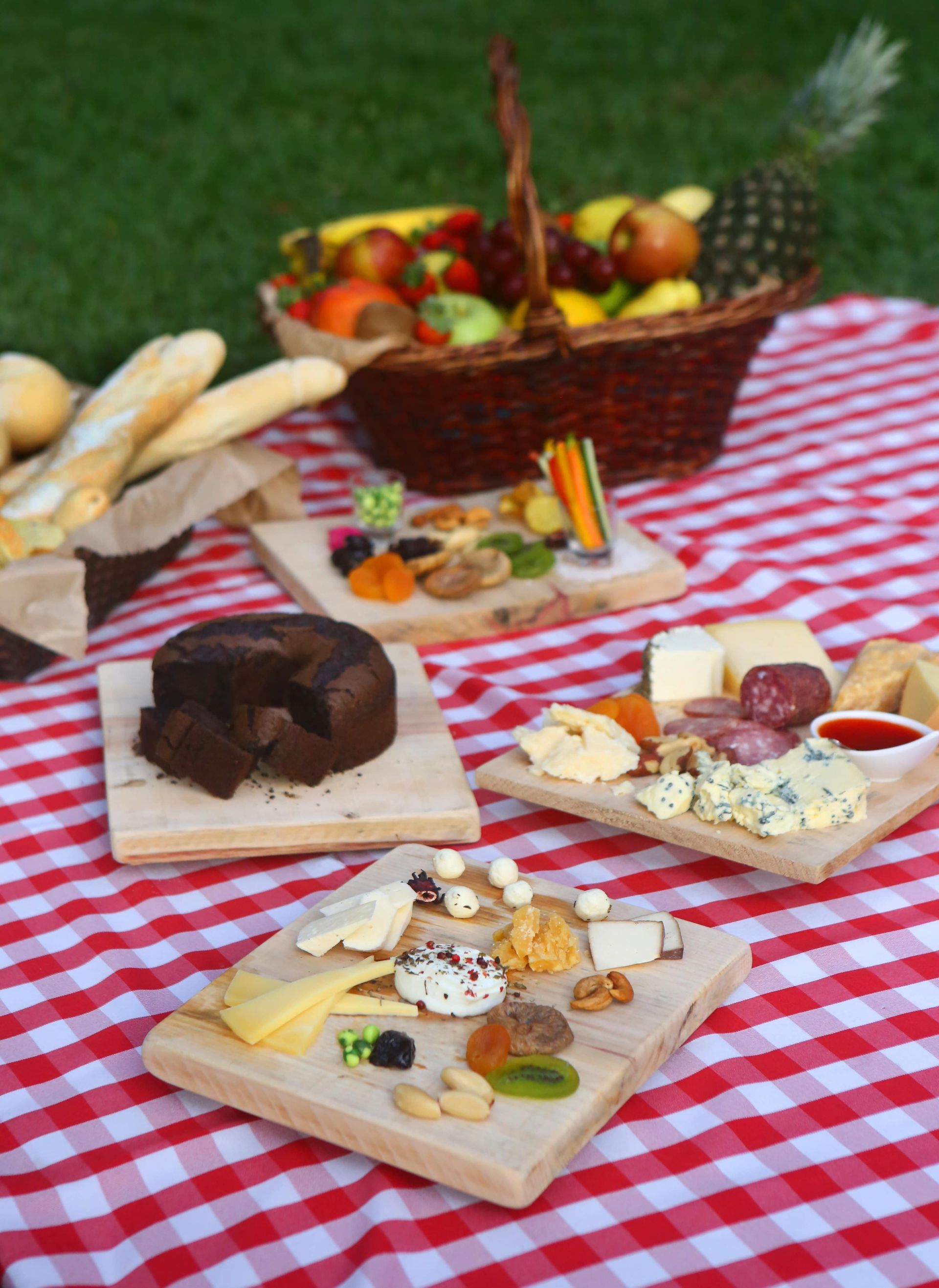 Mesa de piquenique com sobremesas fatiadas, queijo, frutas, pão e uma cesta sobre uma toalha xadrez vermelha e branca.