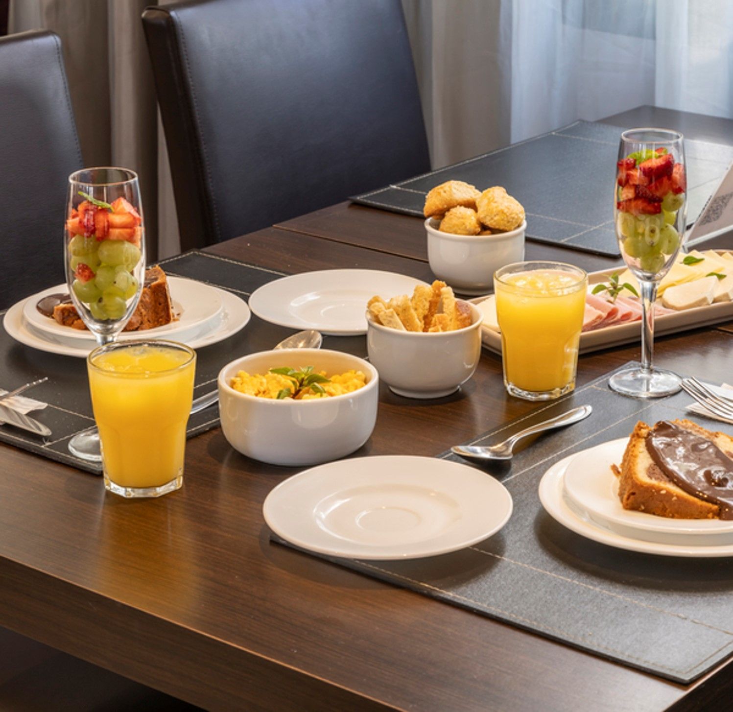 Mesa de jantar com um buffet de café da manhã composto por parfaits de frutas, suco de laranja, doces e tigelas.