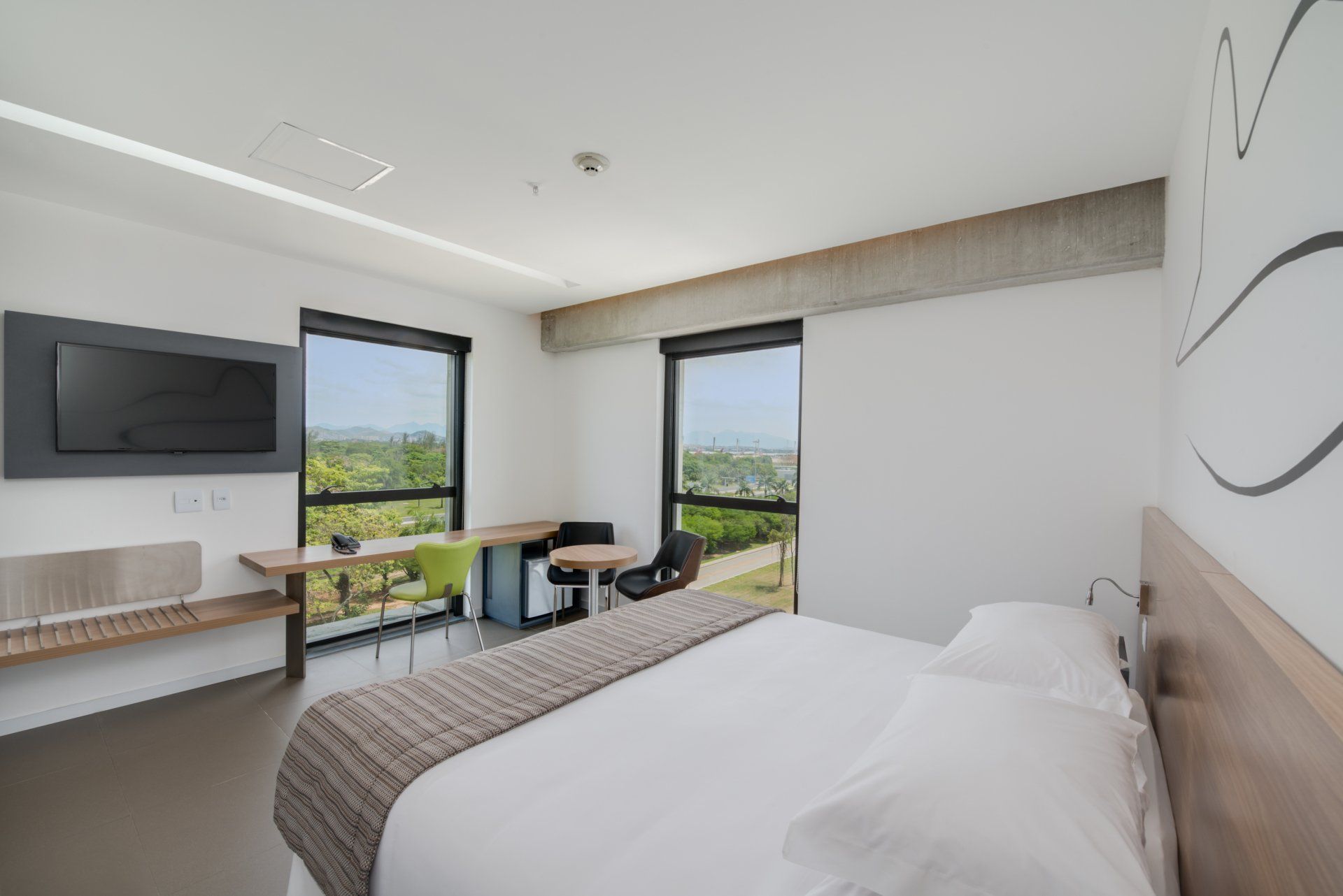 Quarto de hotel moderno com cama, escrivaninha, TV de parede e janelas amplas com vista para a vegetação.
