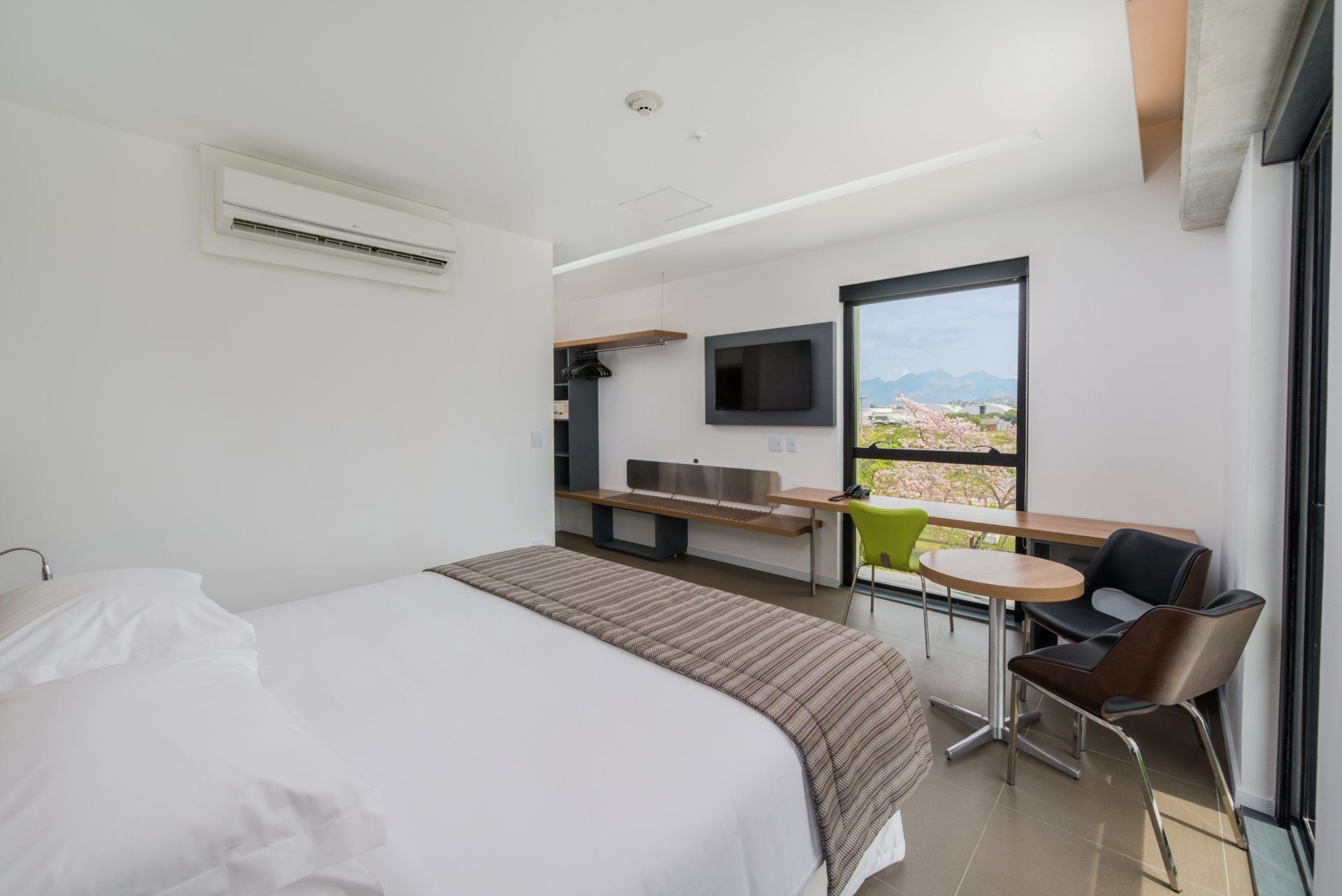 Quarto moderno com cama branca, TV de parede e varanda com vista para as montanhas e para a cidade.