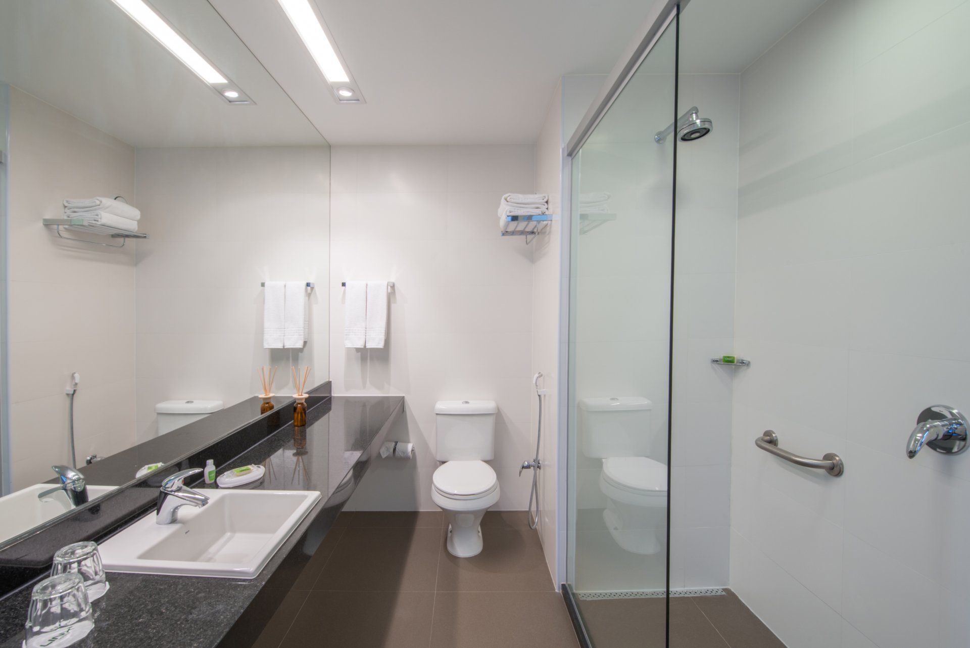 Banheiro público moderno com pias, espelho, vaso sanitário e box de vidro para chuveiro.