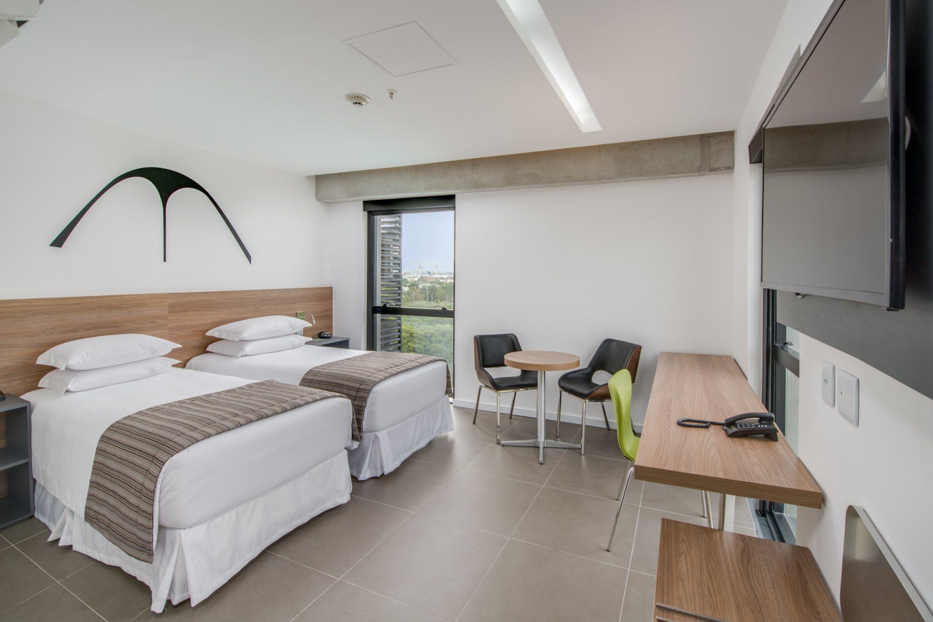Quarto de hotel moderno com duas camas, escrivaninha, cadeiras, TV e vista para a janela.