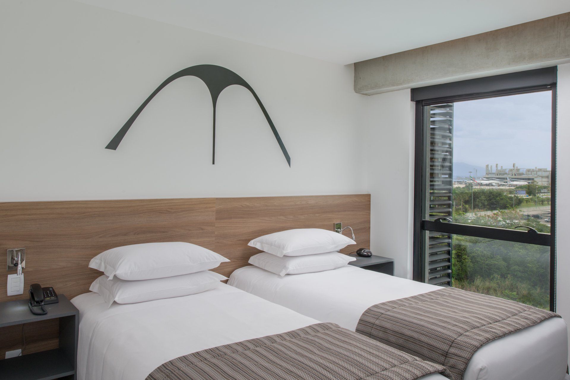 Quarto moderno com duas camas de solteiro, cabeceira de madeira e janela com vista para a cidade.