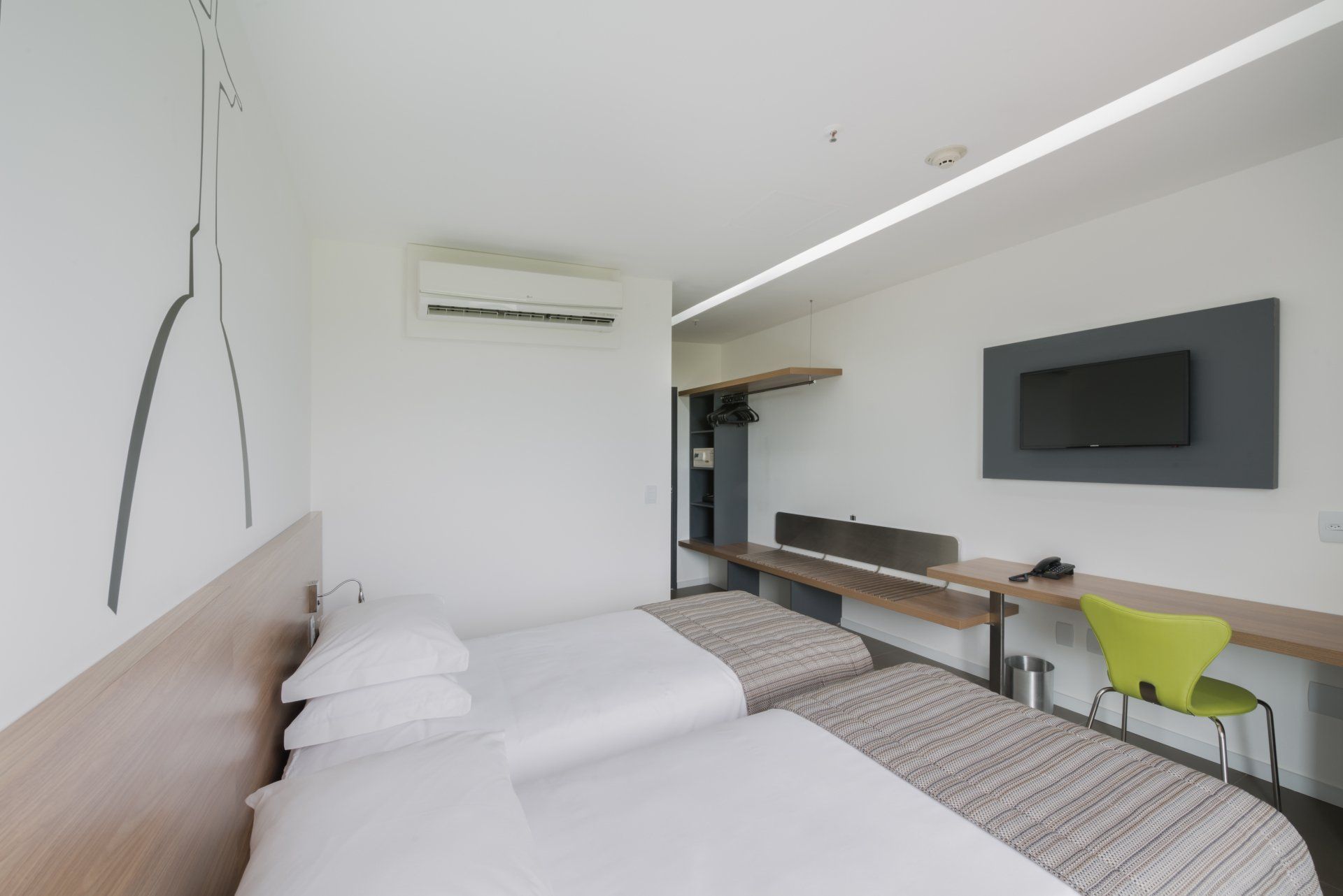 Quarto de hotel luminoso com duas camas, TV de parede, escrivaninha e ar-condicionado.