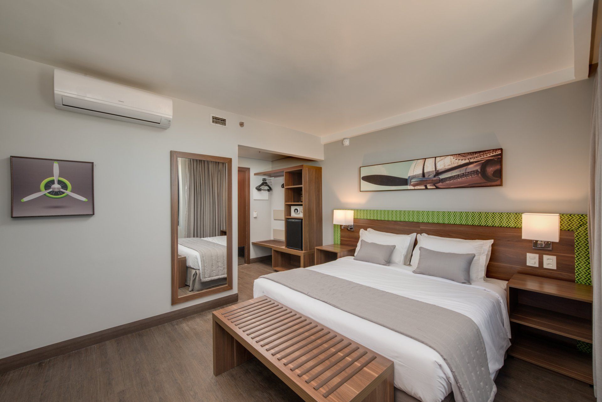 Quarto de hotel moderno com cama king-size, banco, obras de arte na parede, ar-condicionado e iluminação aconchegante ao lado da cama.