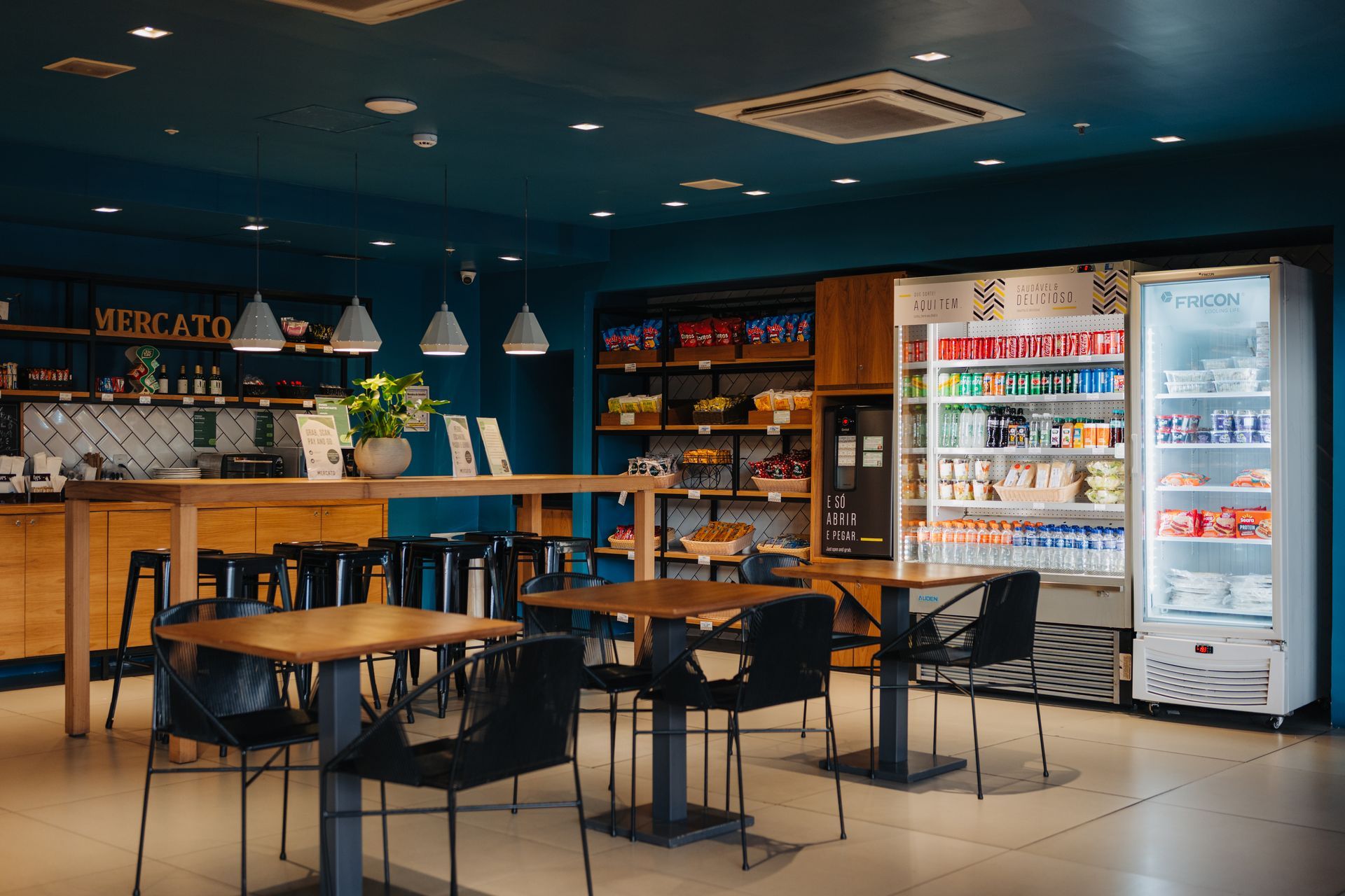 Interior de café moderno com paredes azuis, bancos de bar, mesas e um expositor refrigerado de bebidas.