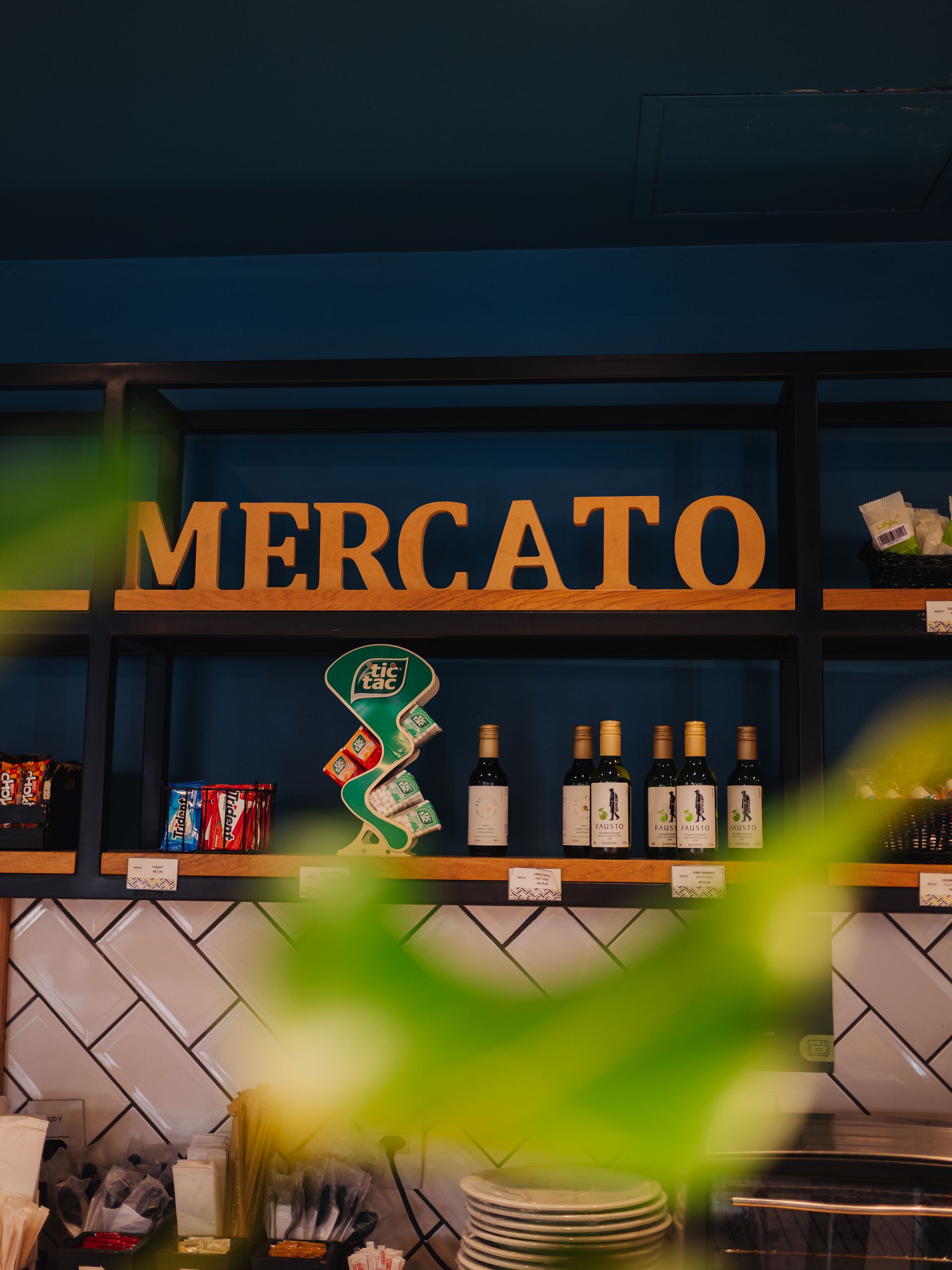 Interior azul de um café com balcão de madeira, banquetas de metal, luminárias pendentes e uma placa com a palavra “MERCATO” atrás do balcão.