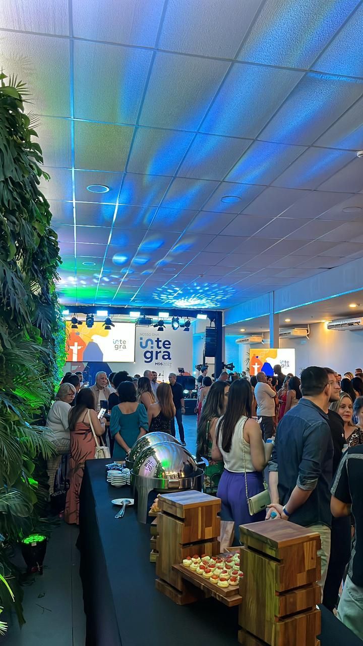 Evento lotado em local fechado, com teto iluminado em azul, pessoas circulando ao lado de um telão e plantas em vasos.