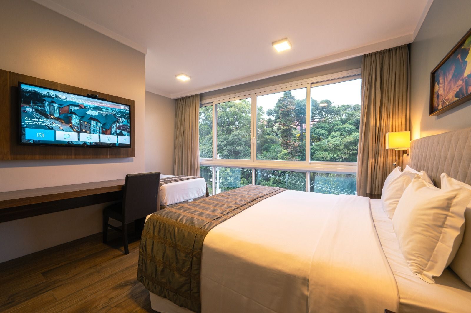 Quarto de hotel moderno com cama, escrivaninha, TV de parede e janela ampla com vista para a vegetação.