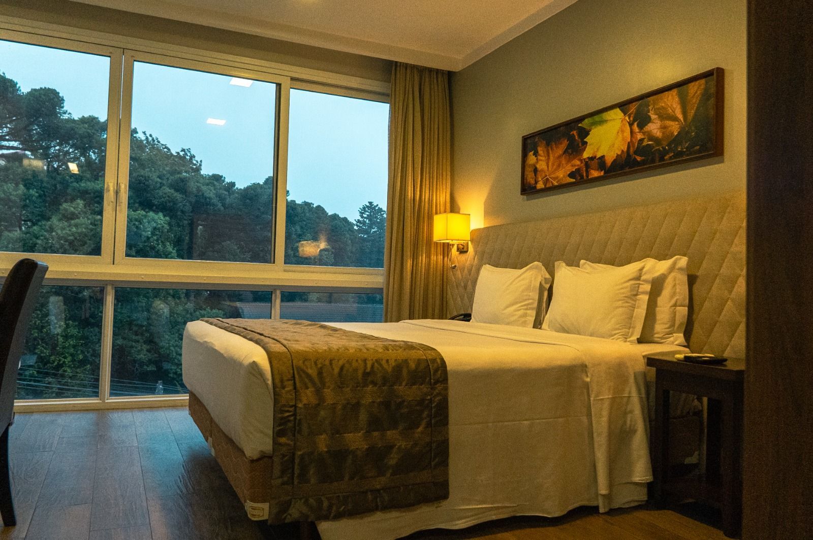 Quarto de hotel aconchegante com cama bem arrumada, luz quente de abajur e janela ampla com vista para as árvores ao entardecer.