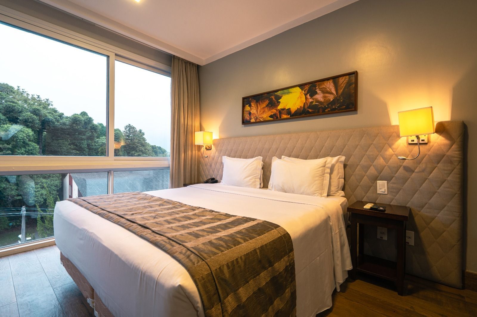 Quarto moderno com cama king-size, luminárias aconchegantes e uma grande janela com vista para a vegetação.