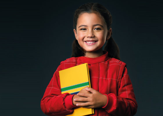 Criança sorridente com suéter vermelho segurando um caderno amarelo contra um fundo escuro.