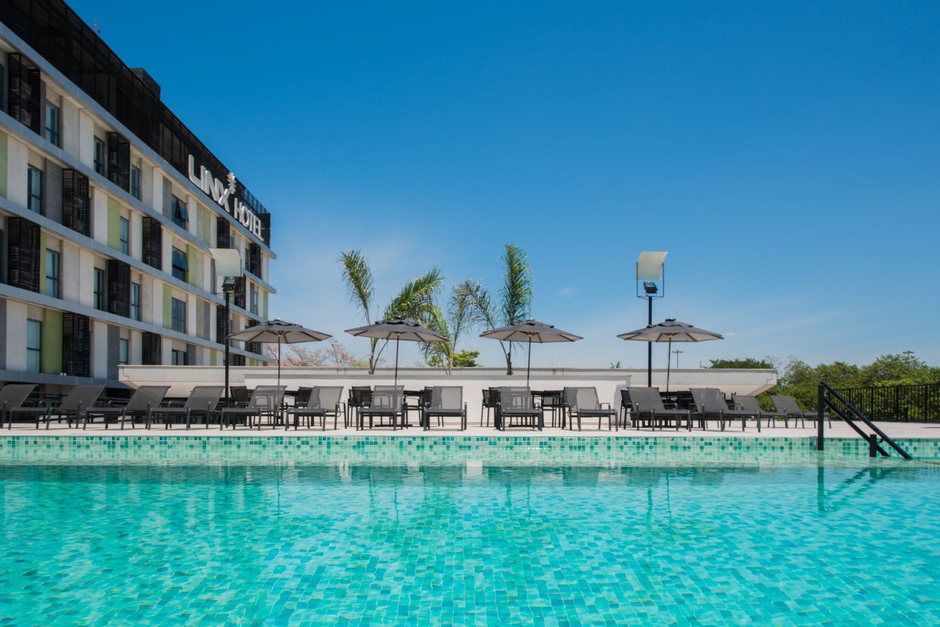 Piscina externa do resort com água turquesa, espreguiçadeiras, guarda-sóis e um prédio do hotel sob um céu azul claro.