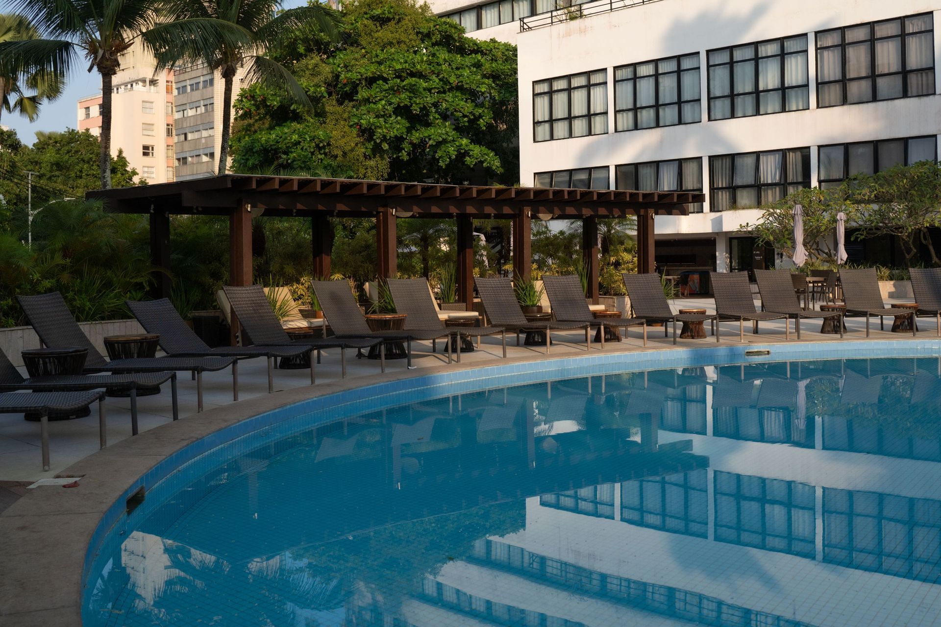 Piscina exterior do hotel com espreguiçadeiras e pérgula ao lado de um edifício moderno.